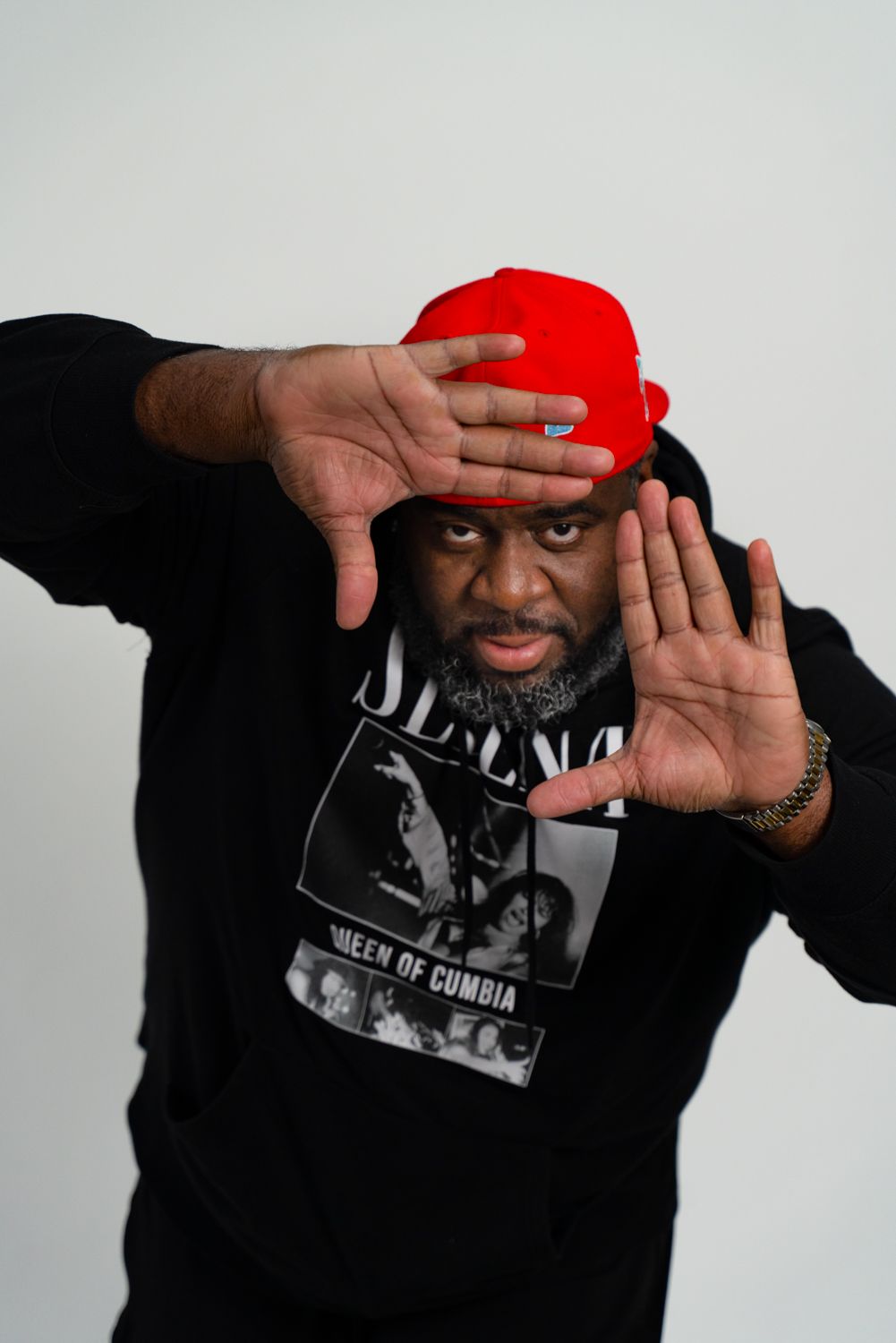 Individual wearing a red cap and black sweatshirt holds hands up in defensive pose against light background.