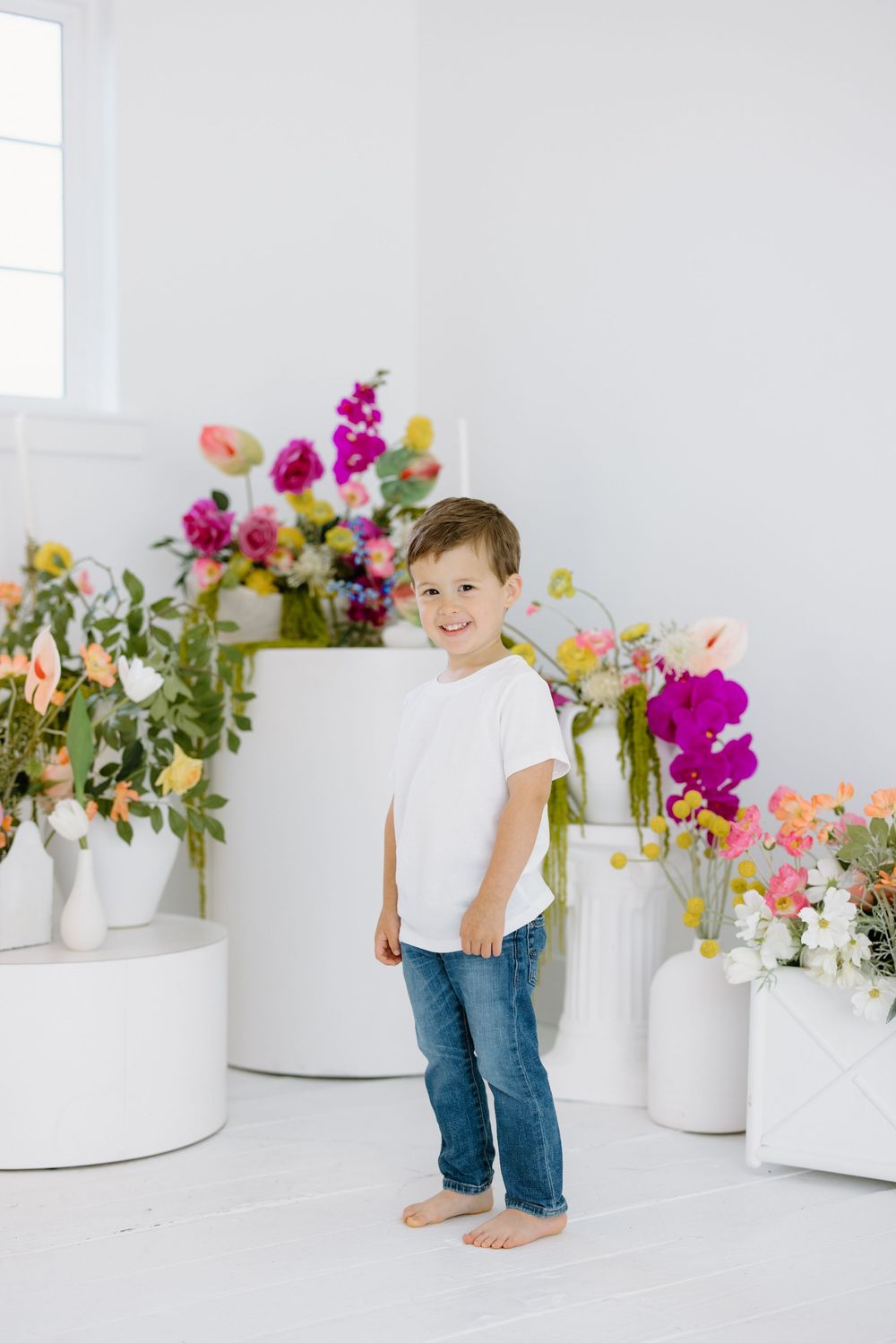 A young barefoot child in jeans and white t-shirt stands among colorful flower arrangements in white vases on a bright set.