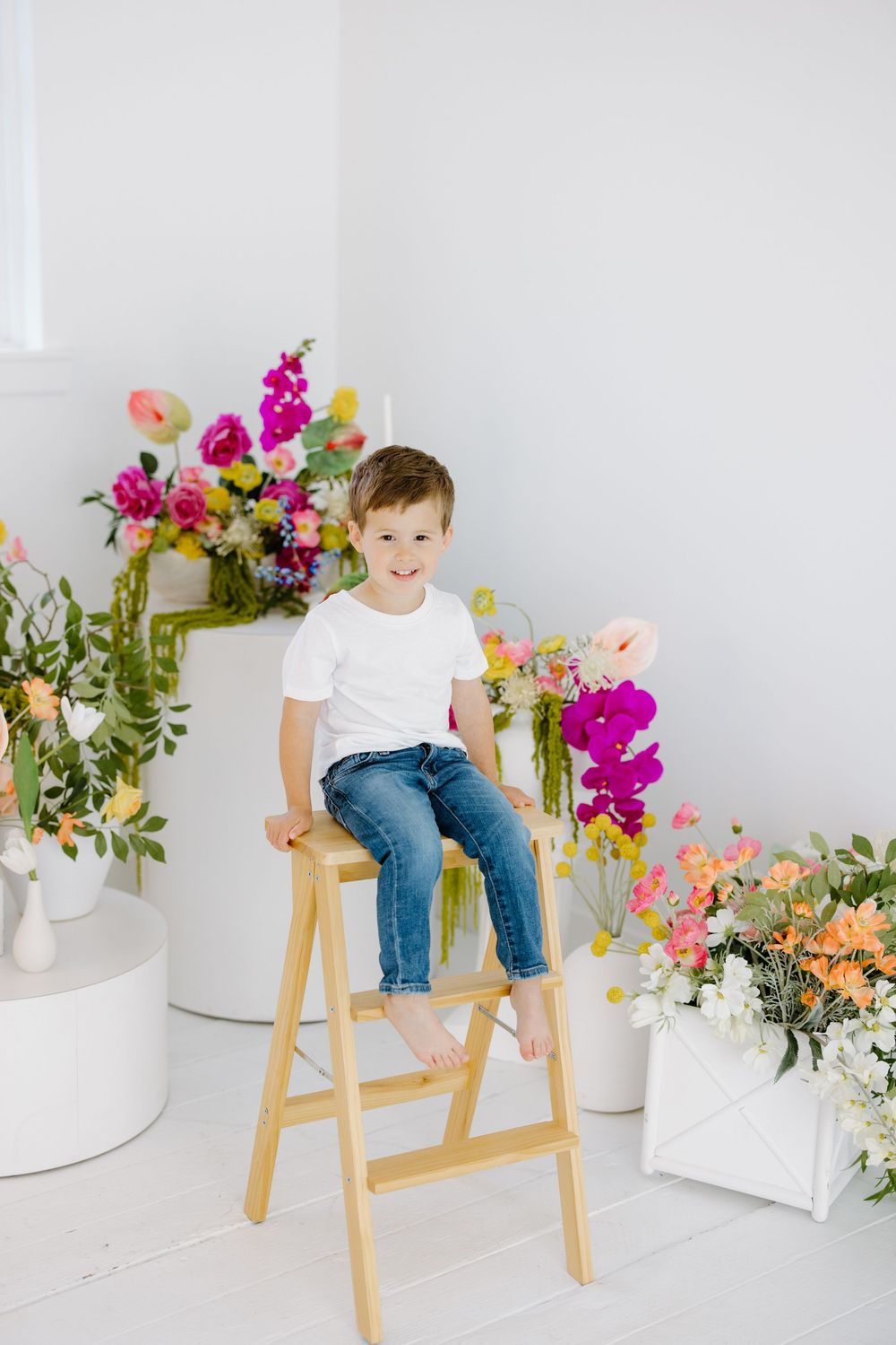 Child sitting on wooden stool surrounded by colorful floral arrangements against white wall.