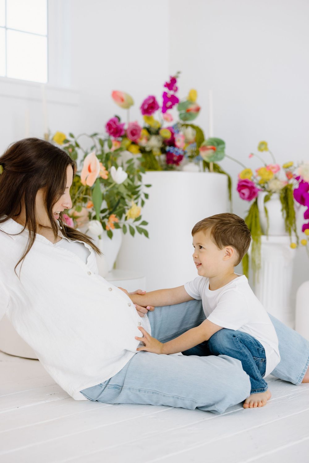 Two people sit together on a white bed with colorful floral arrangements in vases behind them.