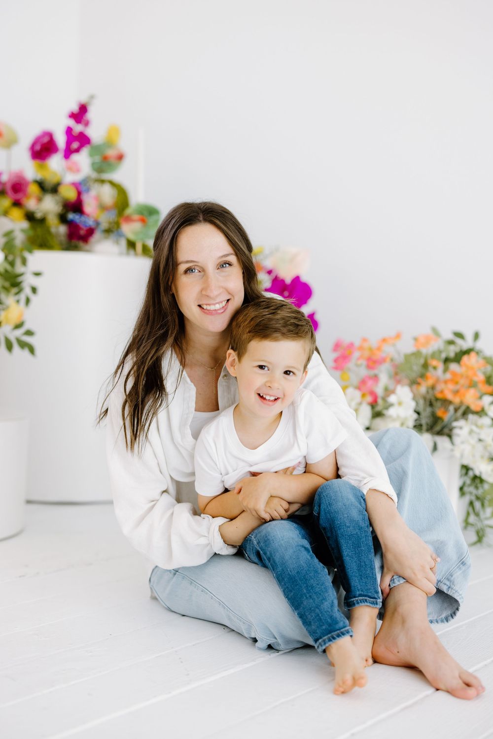 A parent and young child share a tender moment while sitting together on a white floor surrounded by colorful flowers.