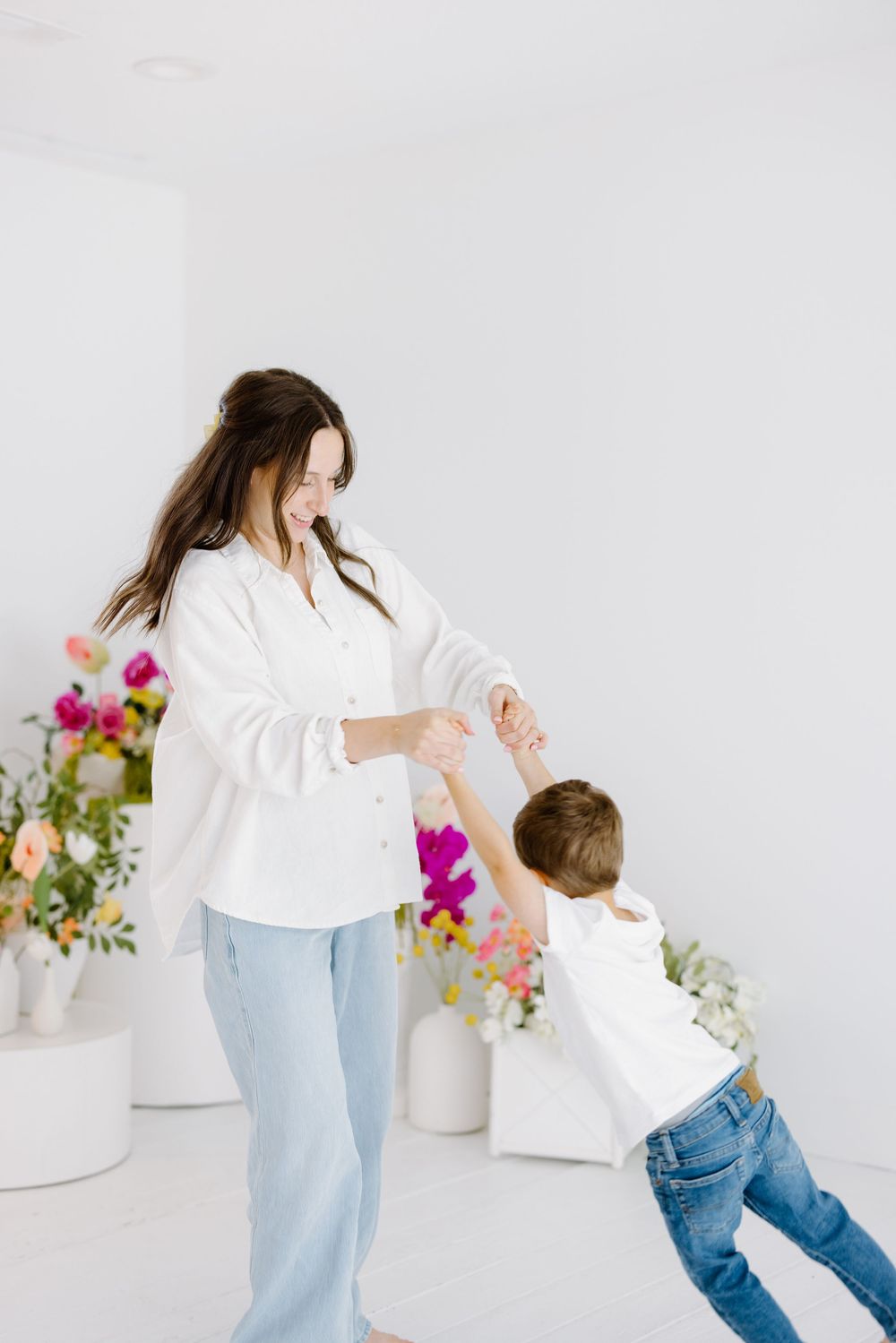 A mother and child in white and denim dance joyfully together against a clean white backdrop with colorful flowers.