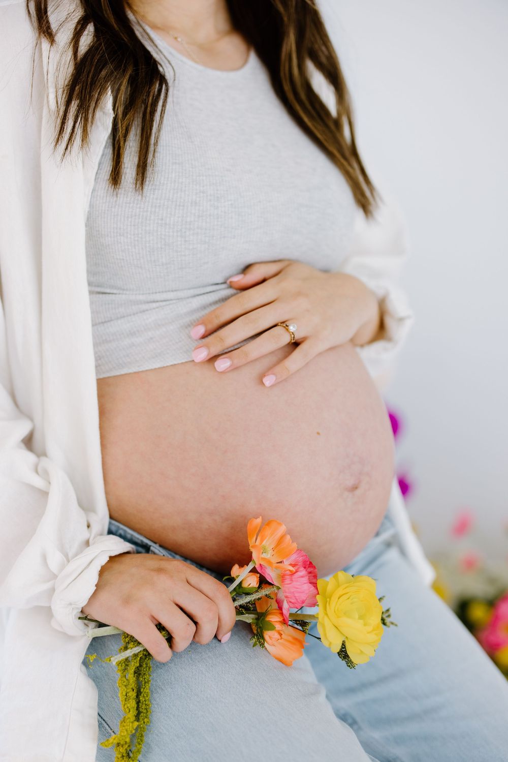 A pregnant woman's belly shown with colorful spring flowers and a white cardigan against a light background.