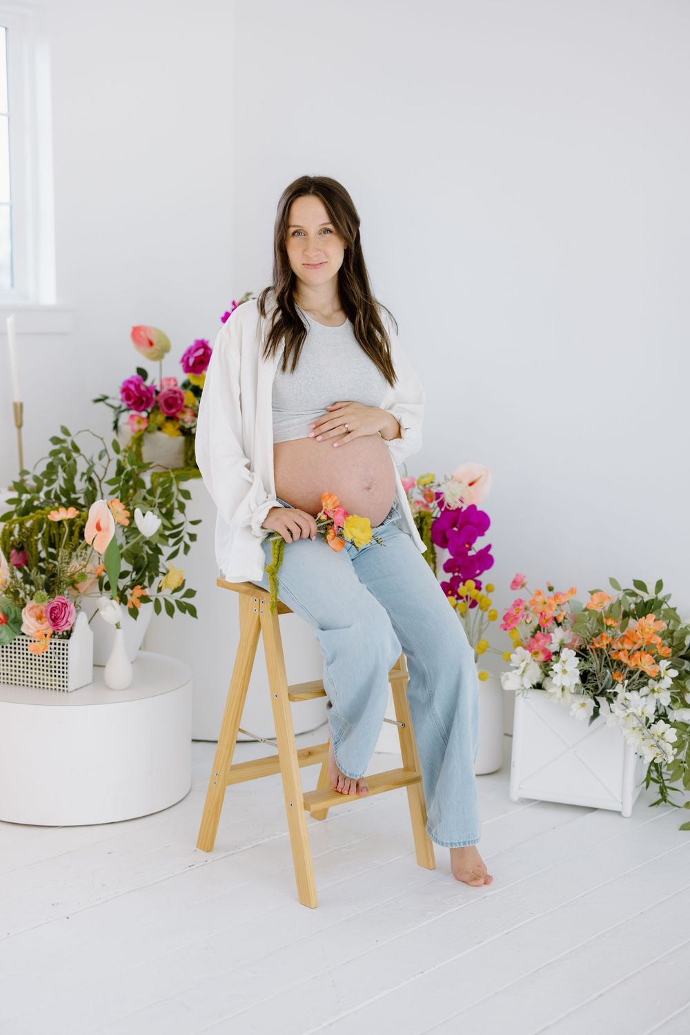 Pregnant woman in white top and jeans sits on wooden stool surrounded by colorful floral arrangements in bright studio setting.