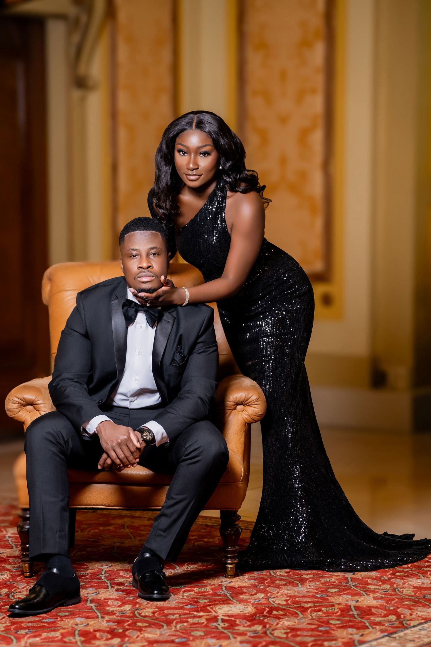Glamorous couple in formal attire pose elegantly with black sequin dress and tuxedo on vintage leather chair.