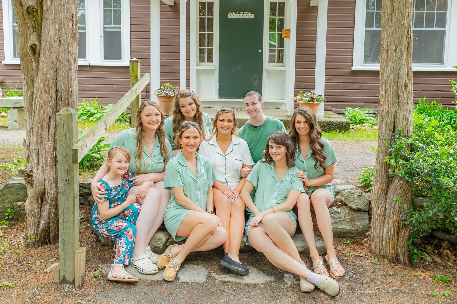 A group of people wearing coordinated mint green outfits sit together on rustic wooden steps in front of a brown house.