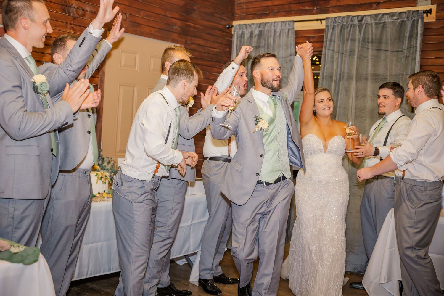 Wedding party wearing matching gray suits and dresses share champagne toasts during celebration at rustic venue.