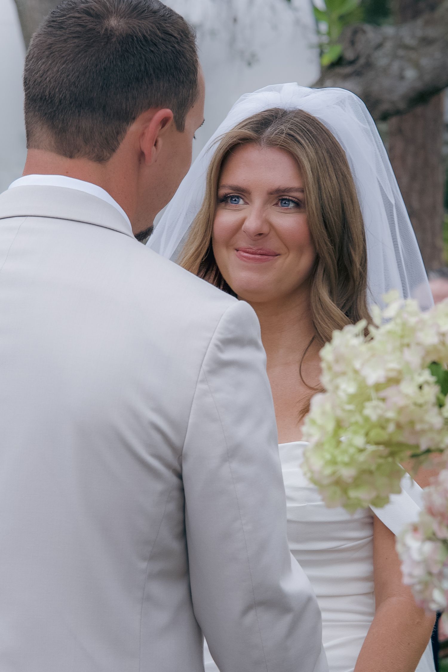 A bride in a white veil smiles while holding a bouquet of white hydrangeas as she faces her groom in a light gray suit.