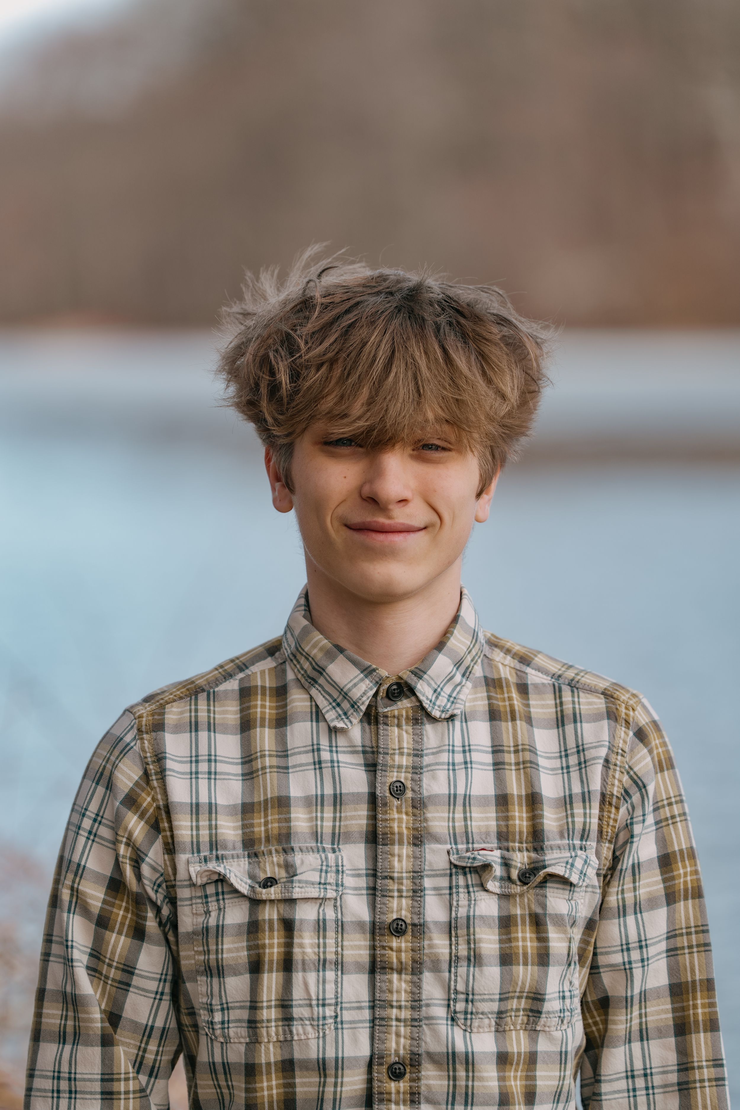 Portrait of someone in a plaid button-up shirt standing outdoors with a gentle smile and tousled hair.