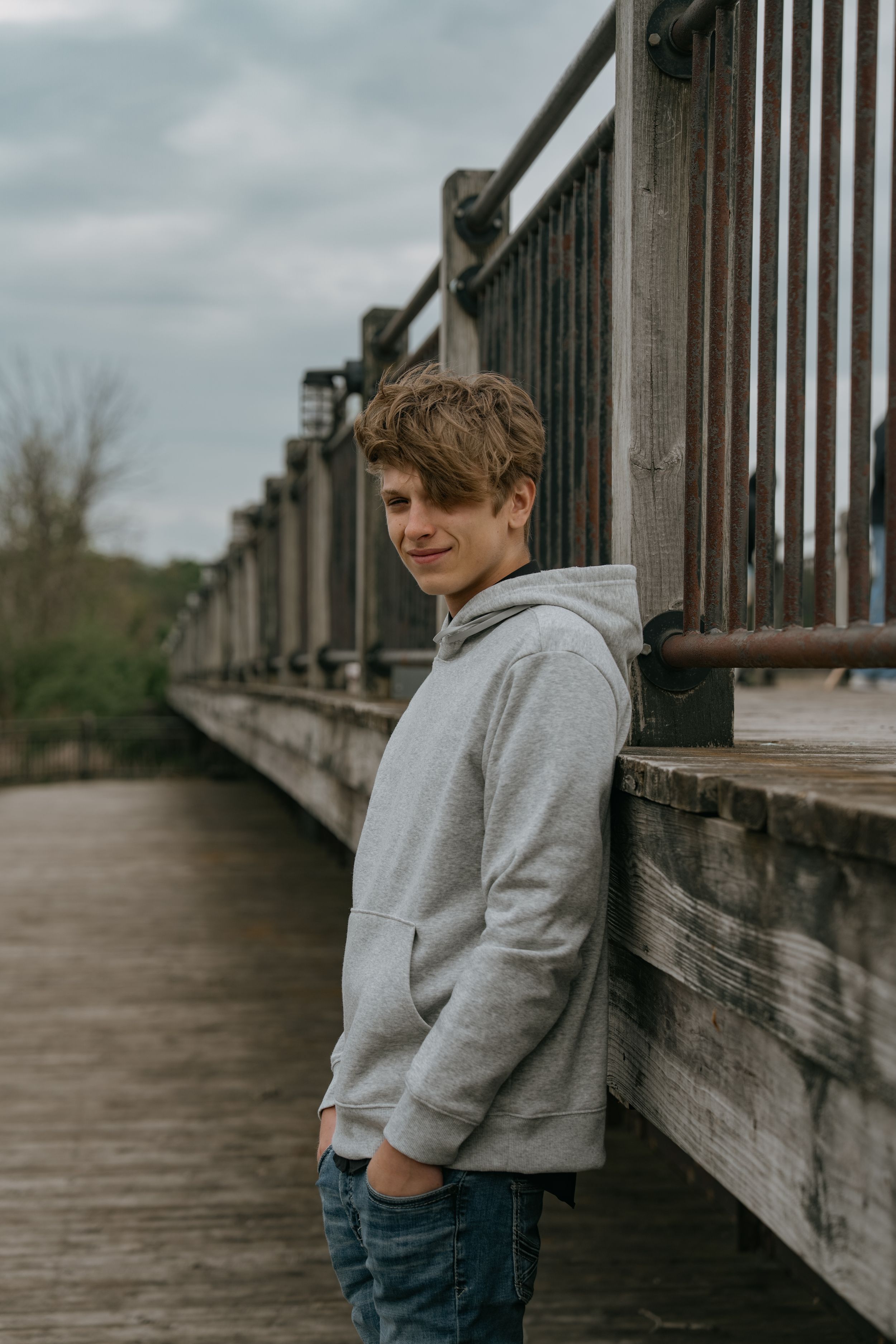 Person in a gray hoodie and jeans standing on a wooden bridge on a cloudy day.