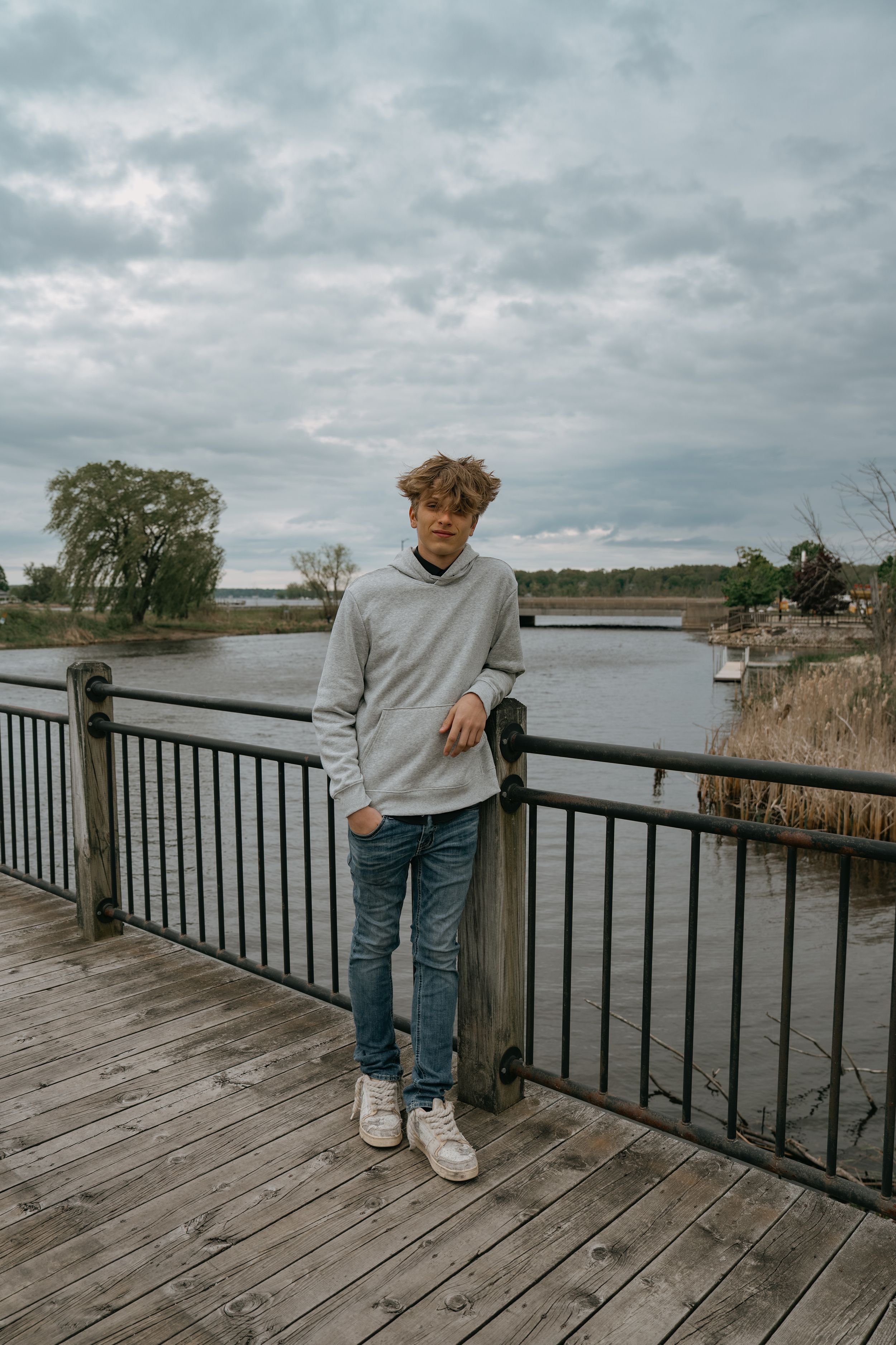 Person in grey sweater and jeans standing on wooden boardwalk overlooking tranquil lake on cloudy day.