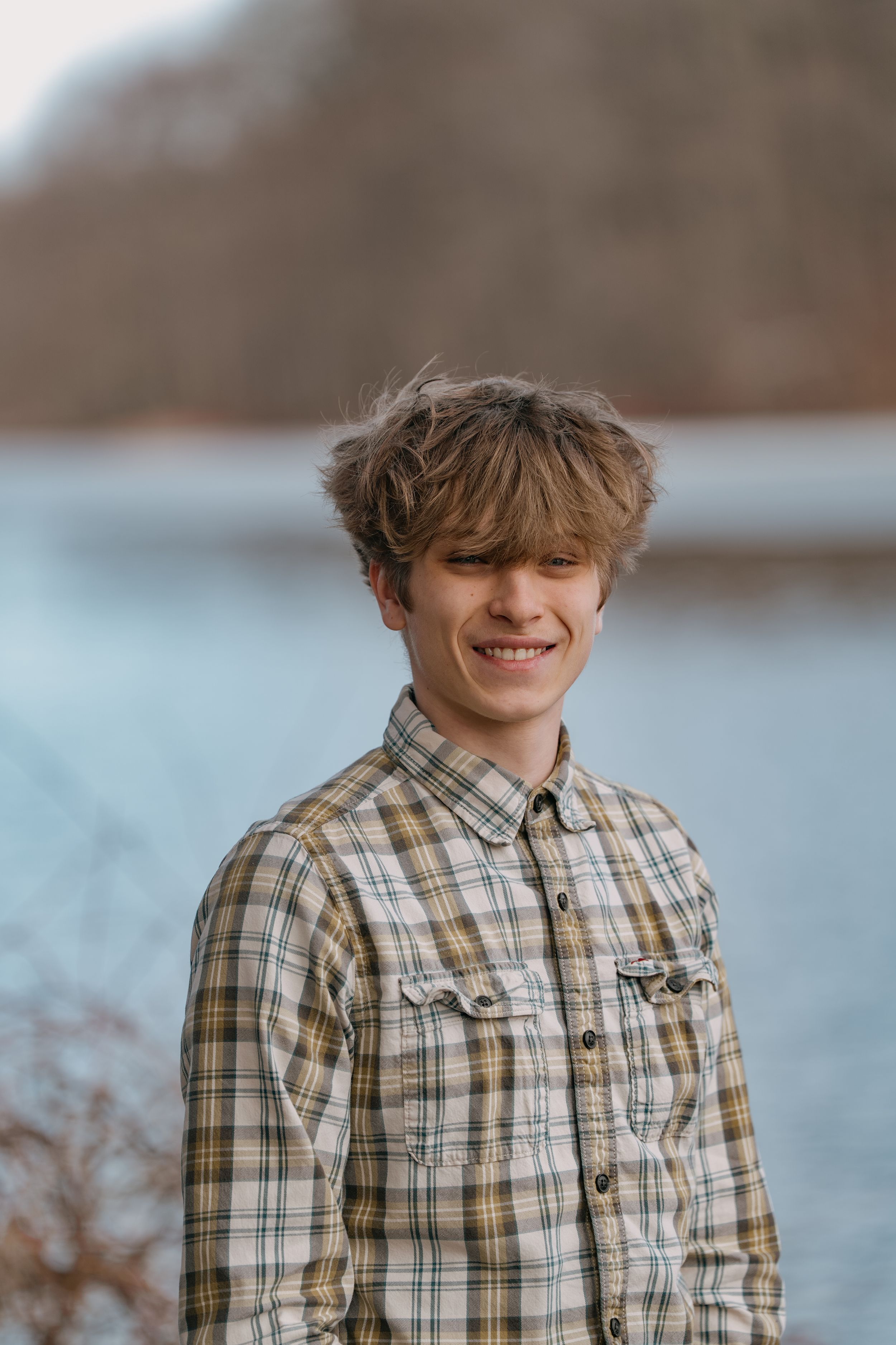 Young person in plaid button-up shirt smiles outdoors with blurred nature background.