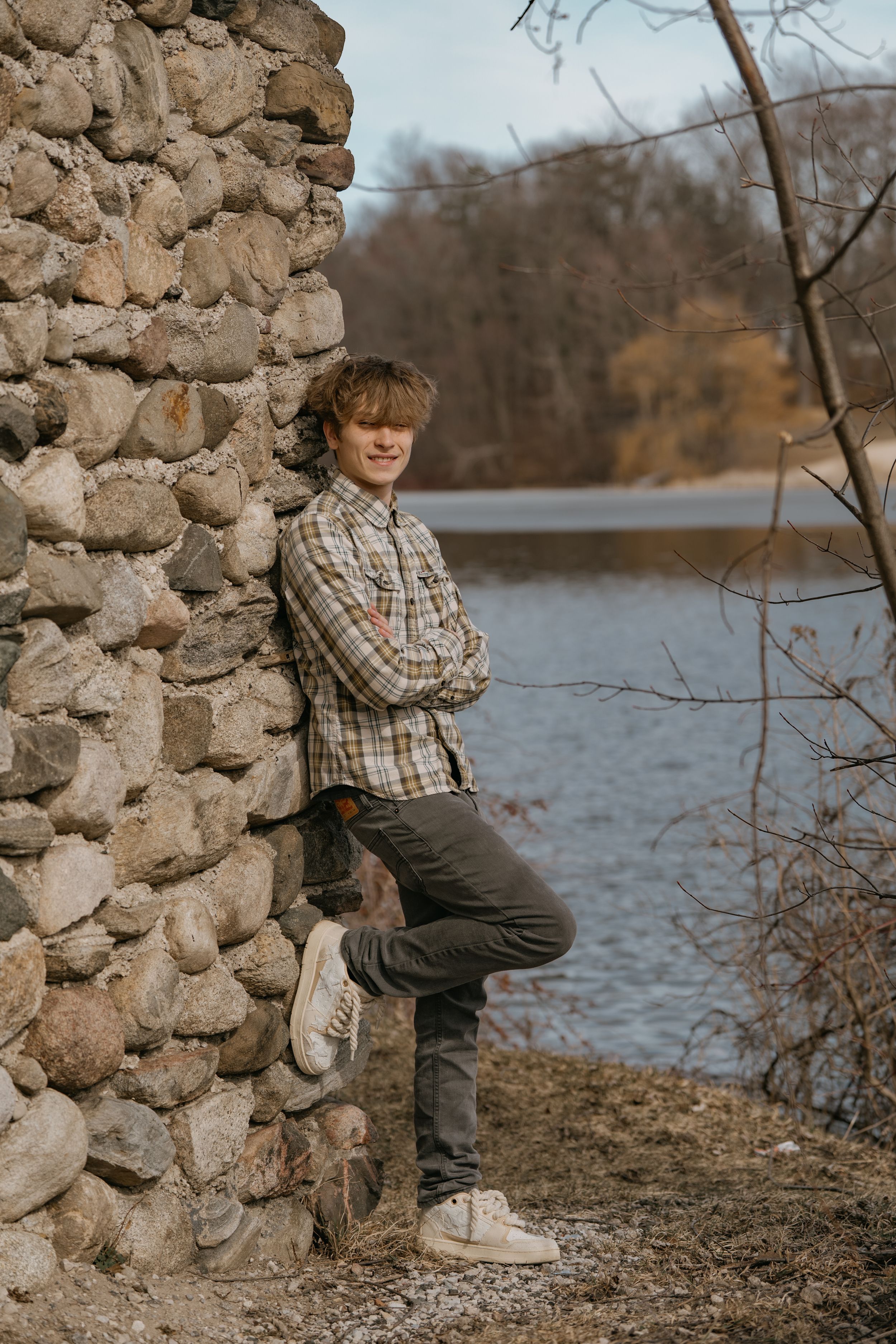 A person in a plaid shirt leans against a stone wall beside a lake on a cloudy autumn day.