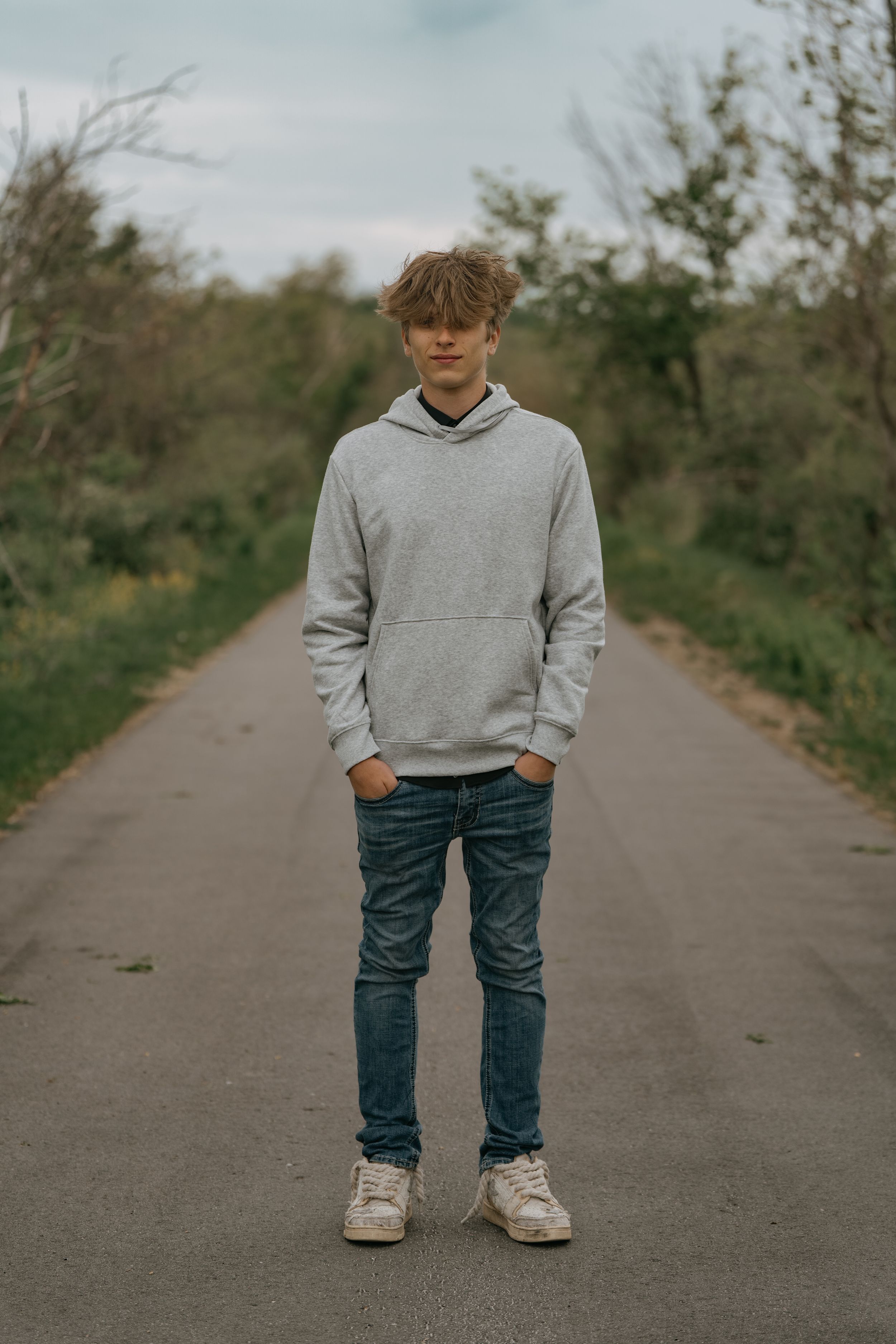 A figure wearing a gray hoodie and blue jeans stands centered on a path with trees in the background.