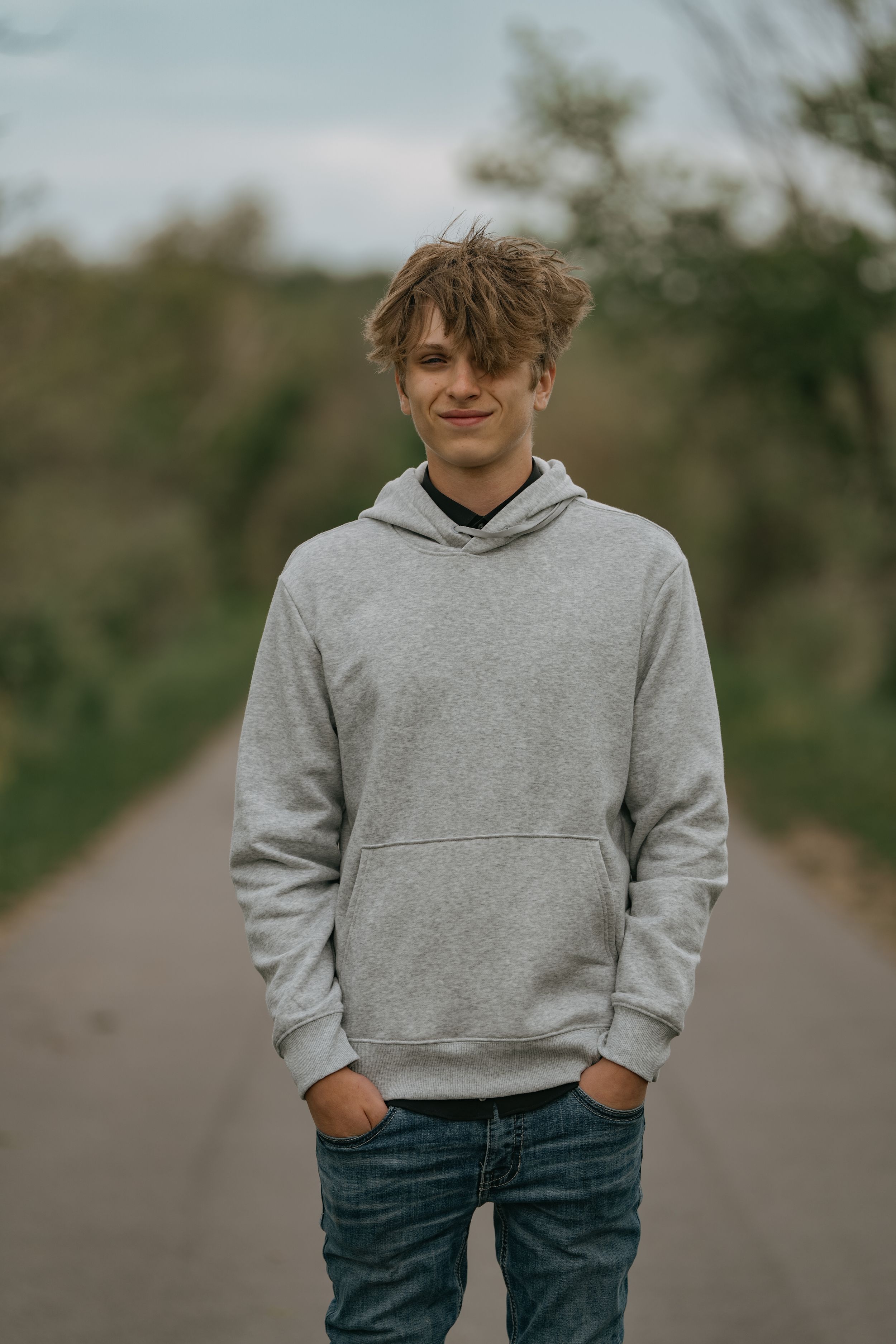 A young person wearing a gray hoodie and jeans stands casually with hands in pockets on a tree-lined path.