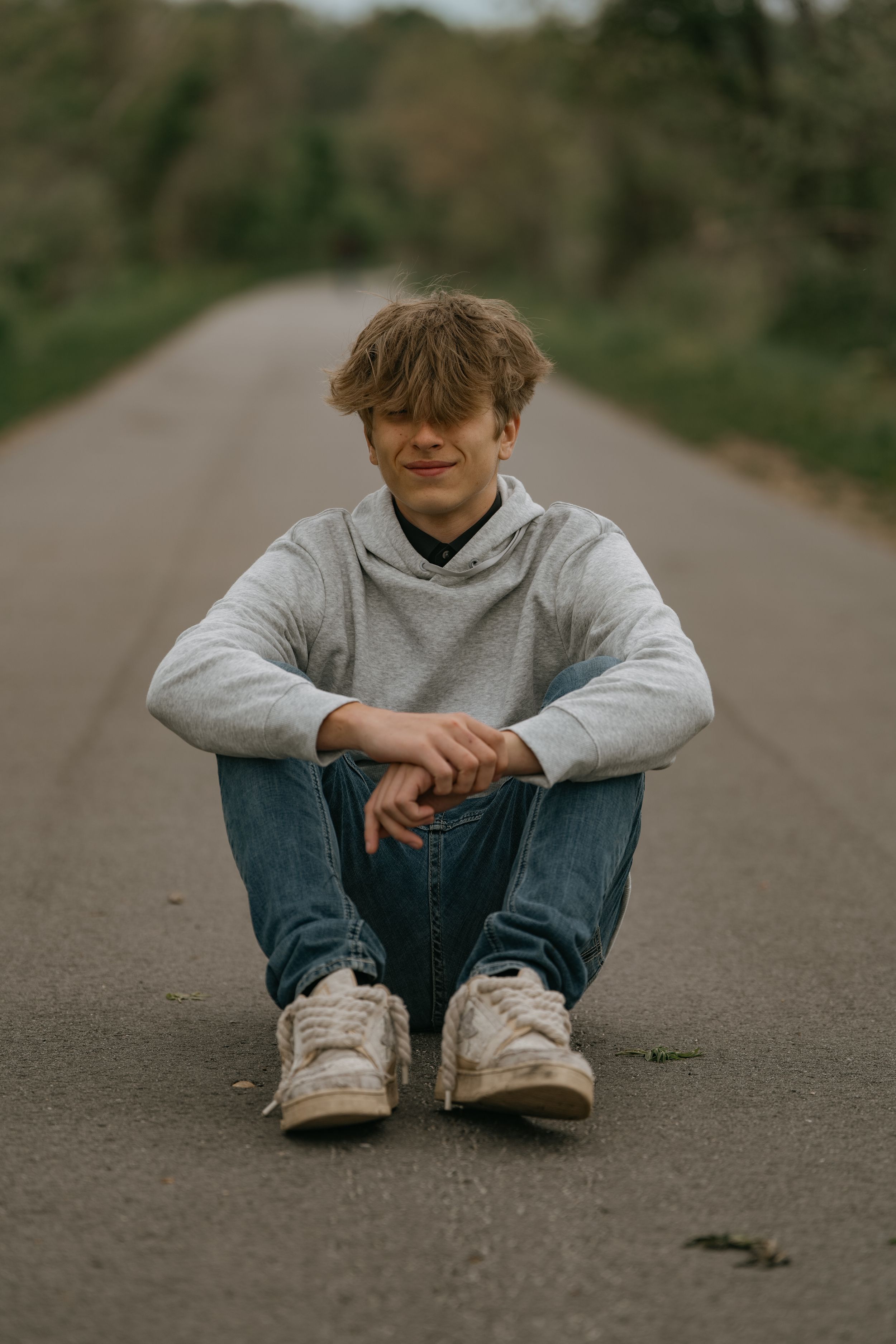 Casual portrait of someone sitting cross-legged on an empty road wearing a gray hoodie and jeans.