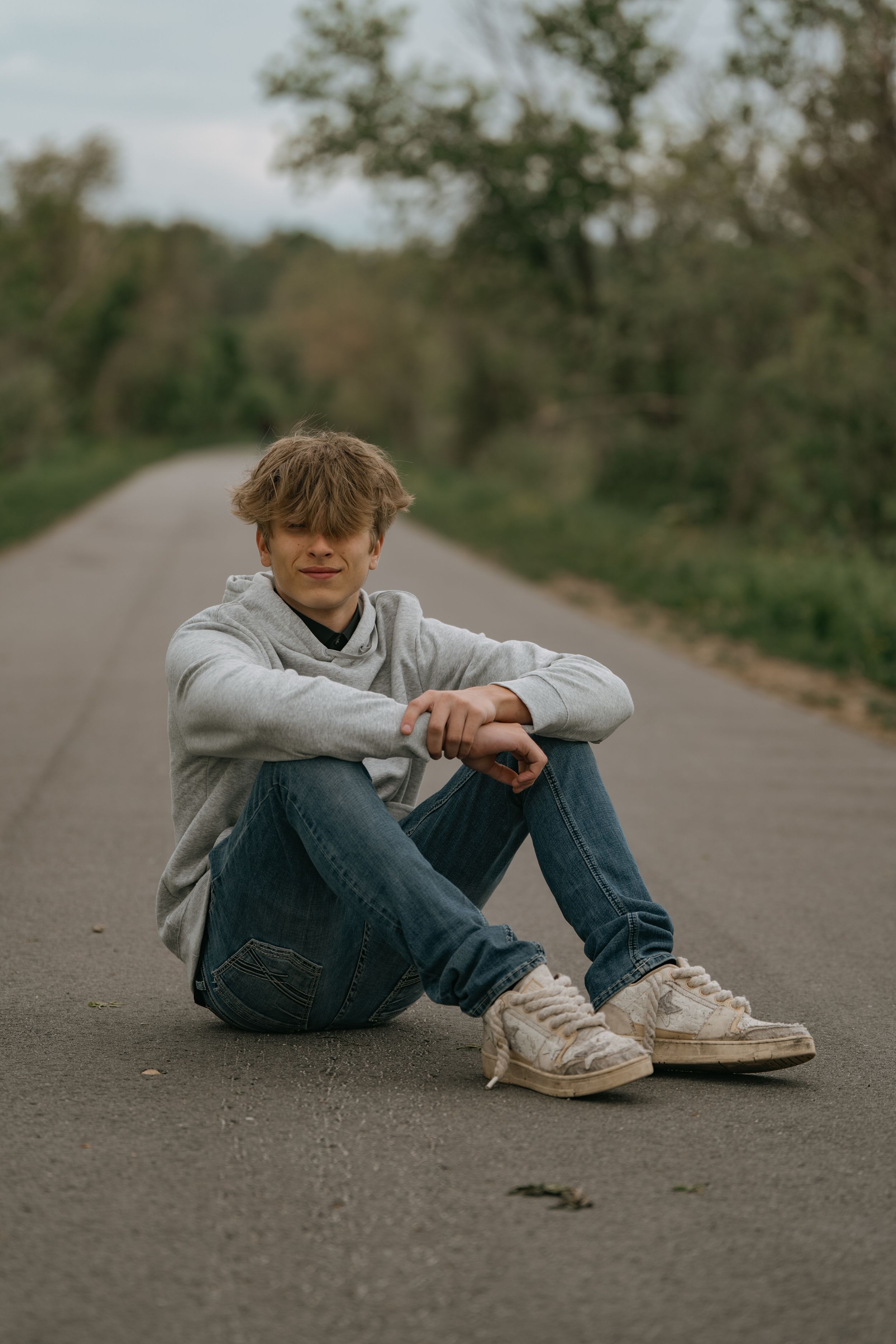 Casual photo of someone sitting on an empty road wearing a grey hoodie, jeans and sneakers.