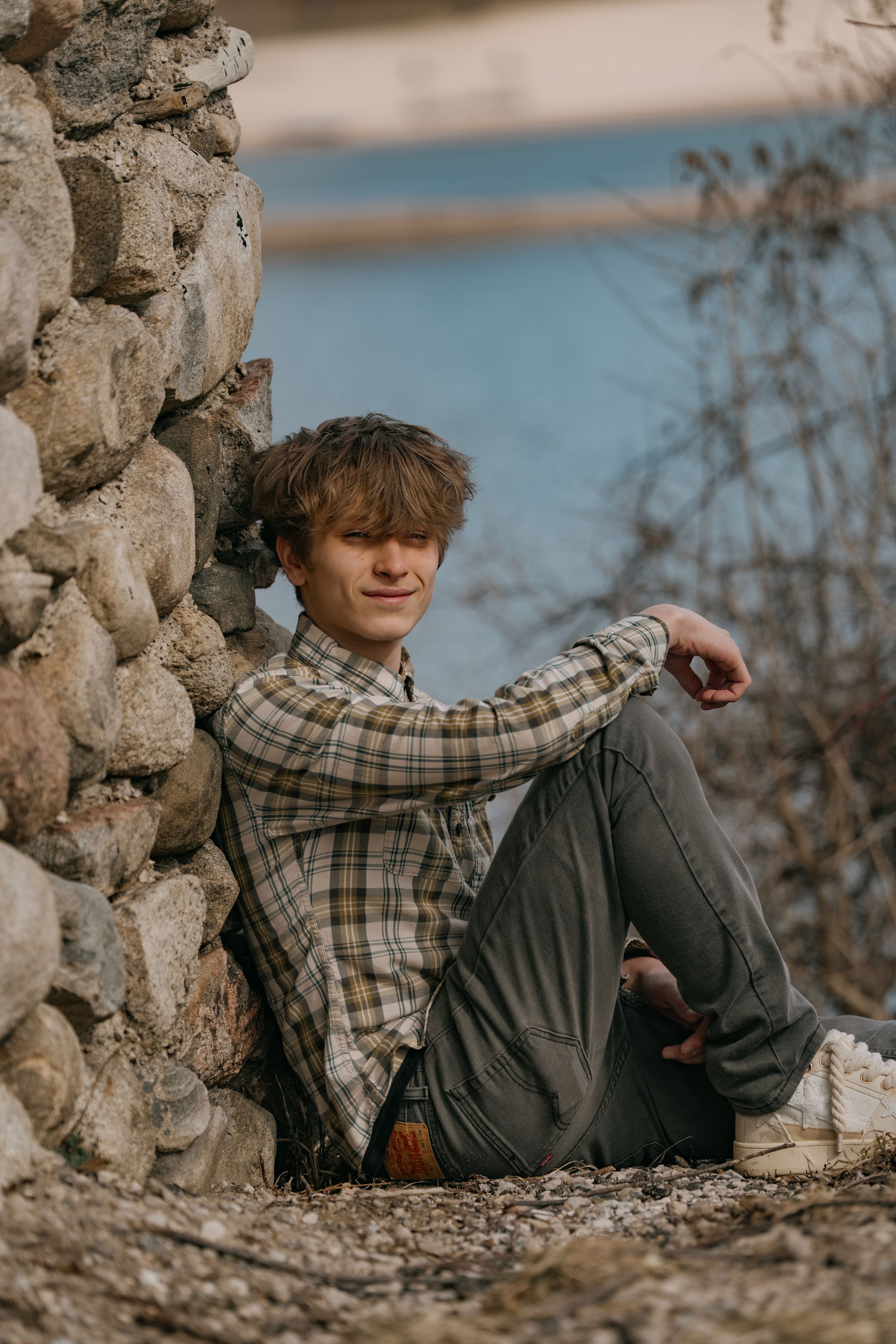 Someone in a plaid shirt and gray pants sits casually against a rustic stone wall during a sunny afternoon.