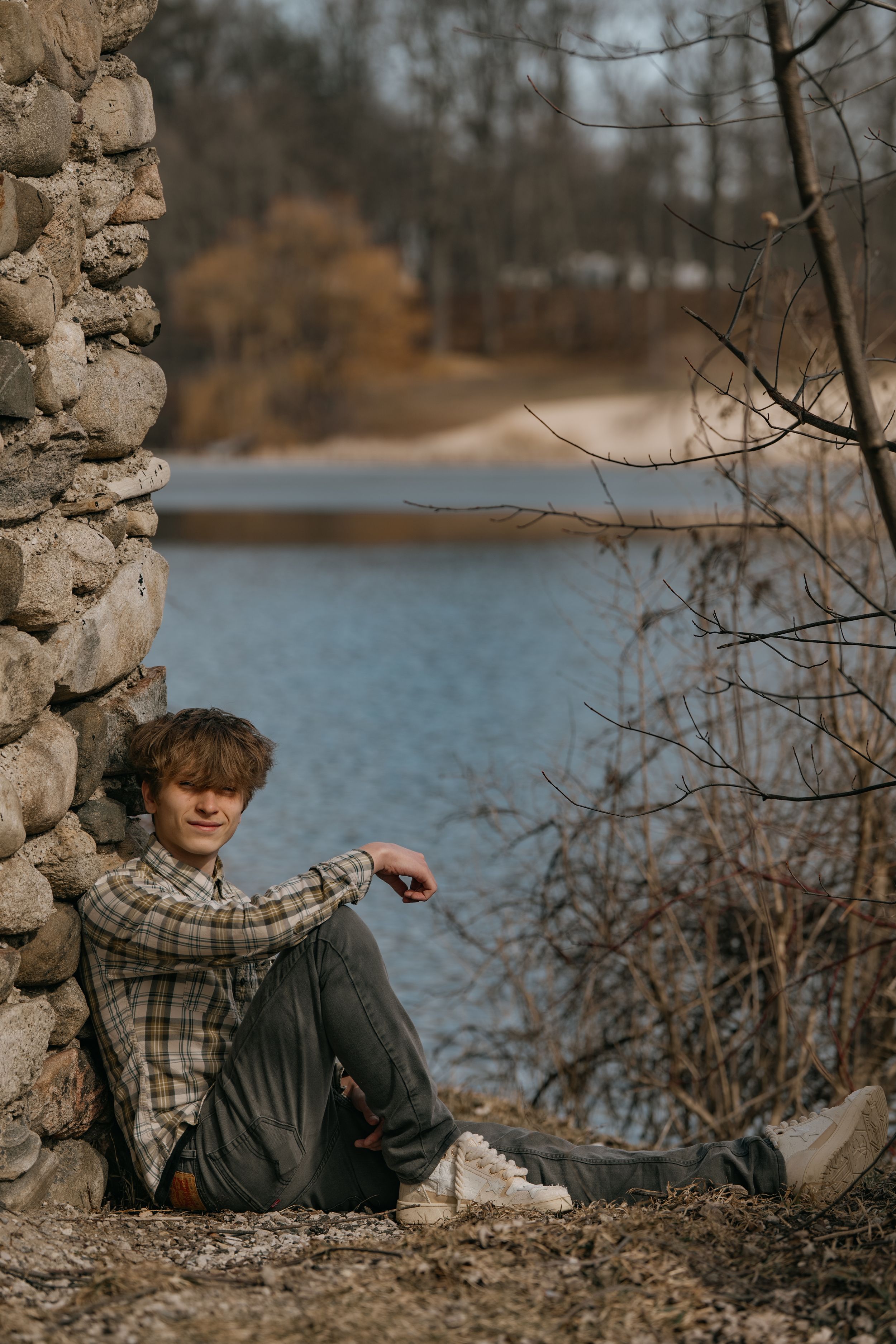 Person in a plaid shirt sits against a stone wall by a serene lake surrounded by autumn trees.