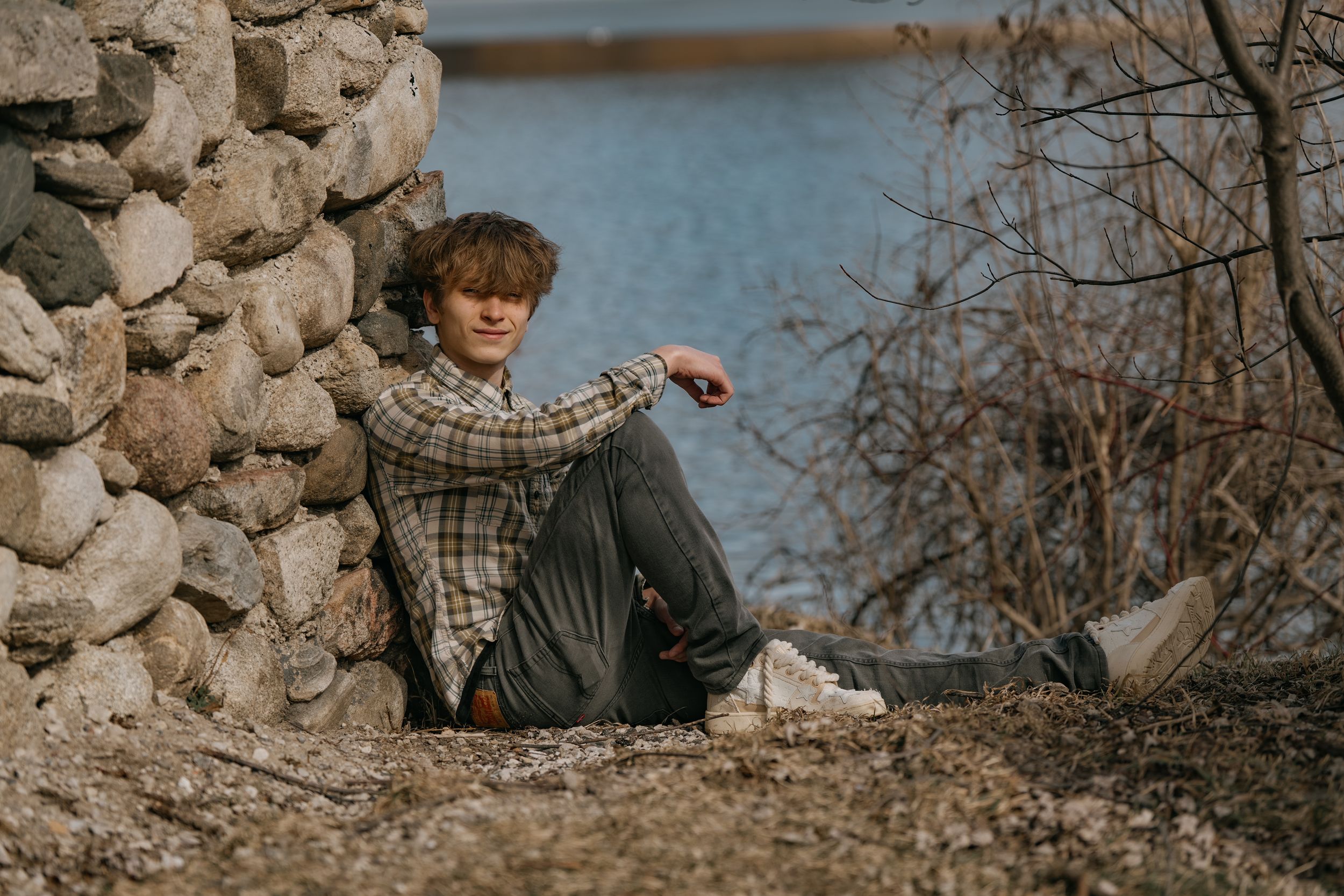 A person sits on the ground beside a stone wall near a lake with bare trees in the background.