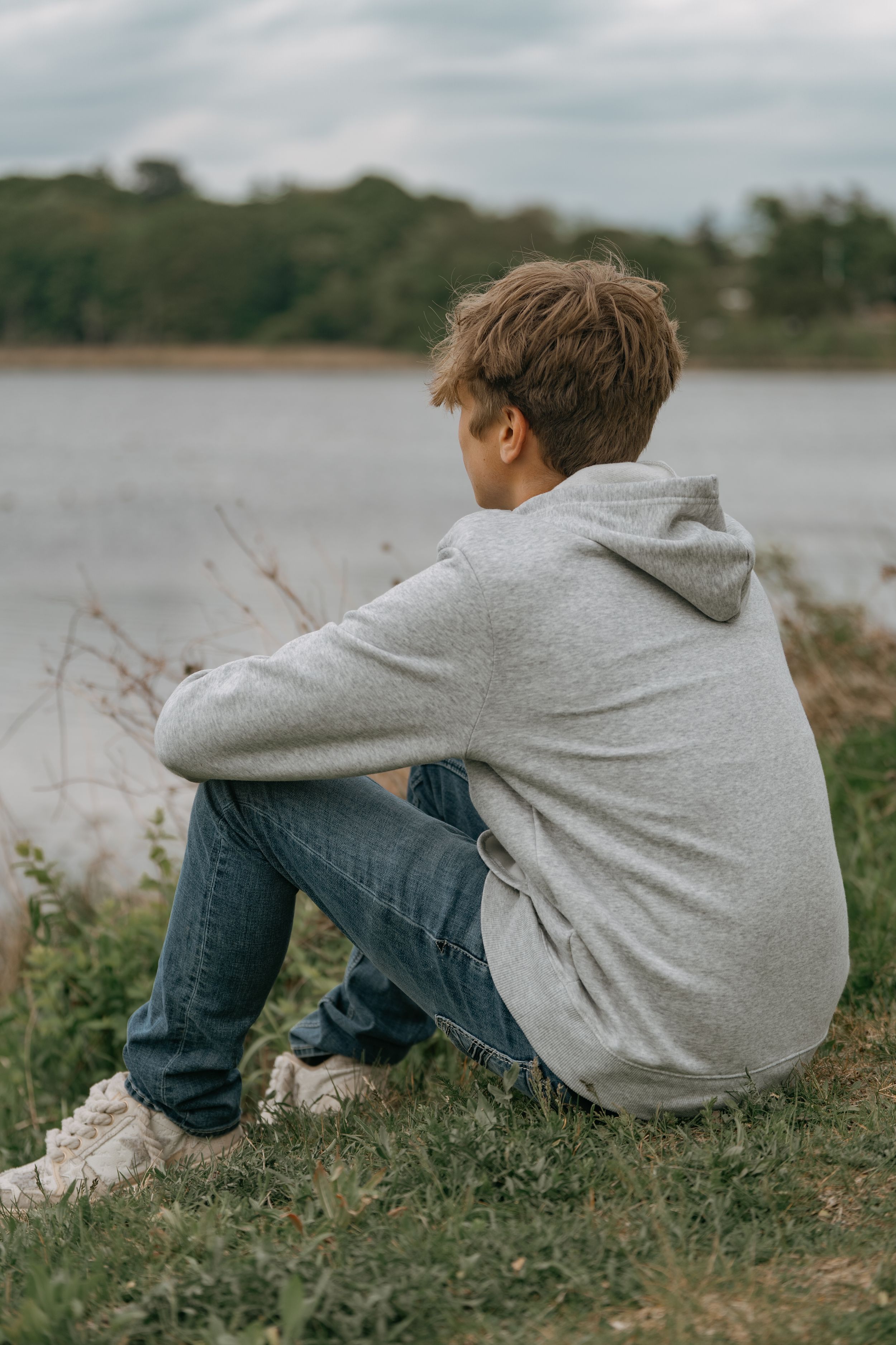 A figure in gray hoodie and jeans sits by a lake shore on an overcast day.