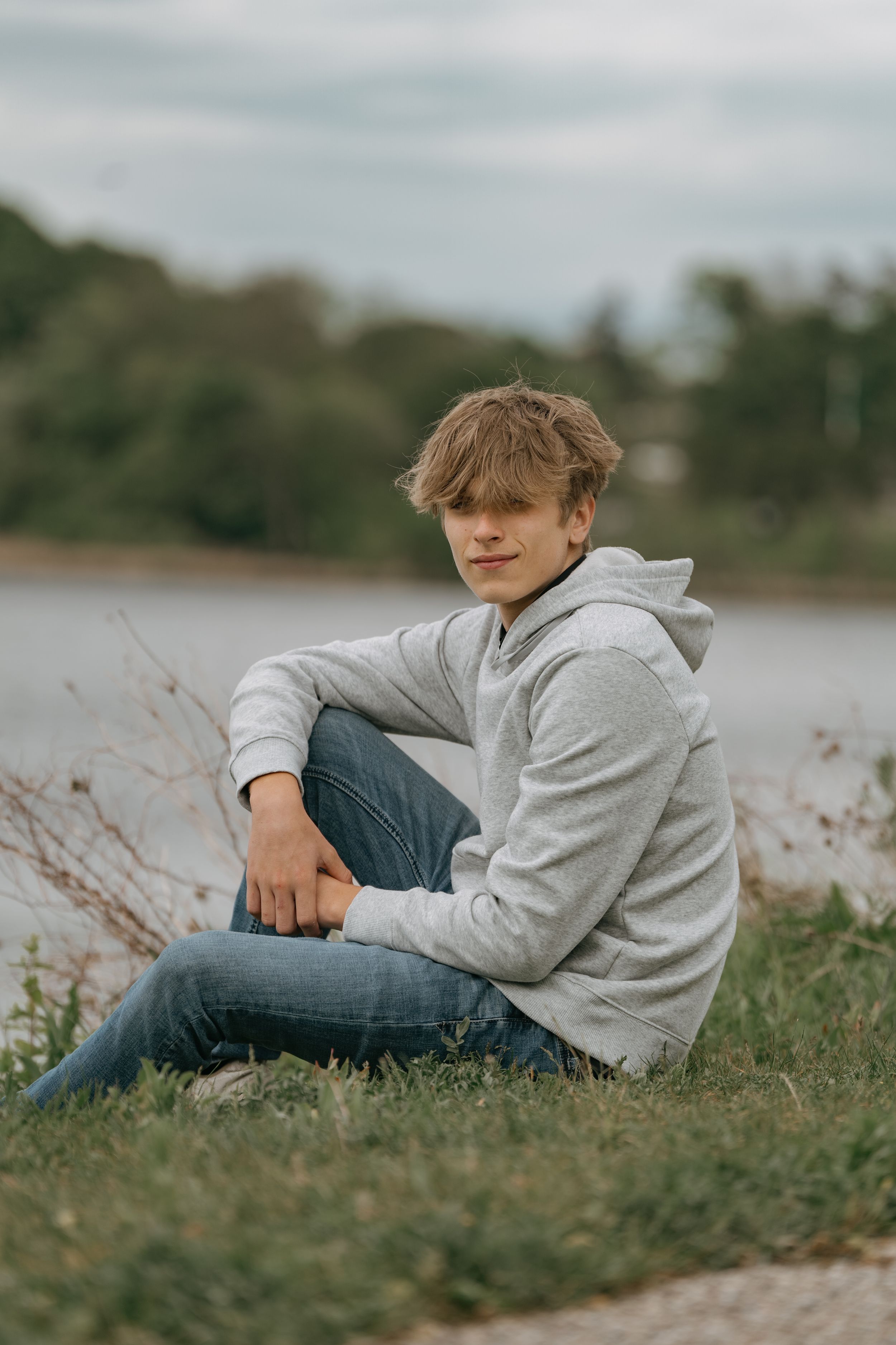 Person in light gray hoodie and jeans sits casually on grassy lakeside bank on an overcast day.
