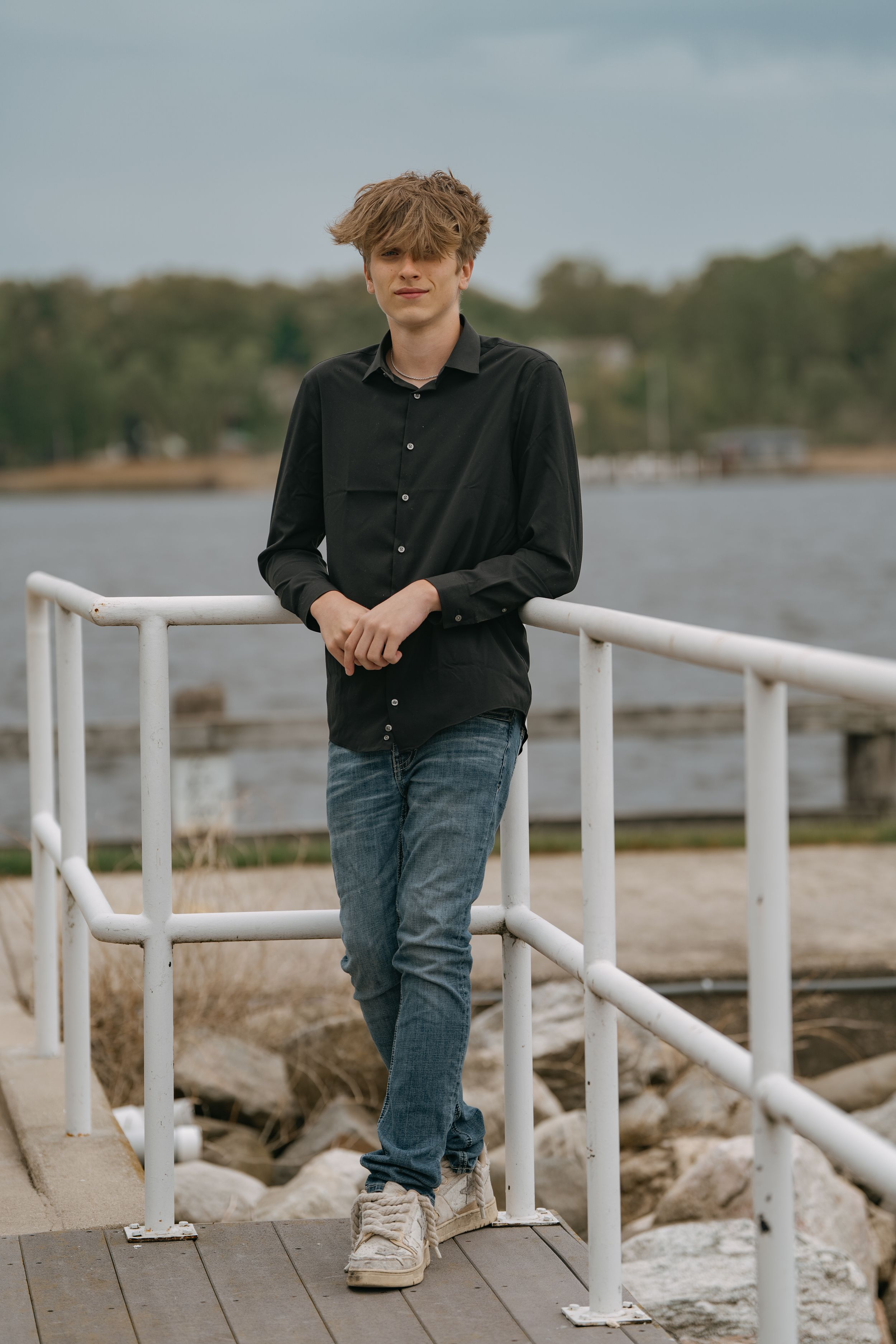 Young person in black button-down shirt and jeans standing on wooden dock by water on cloudy day.