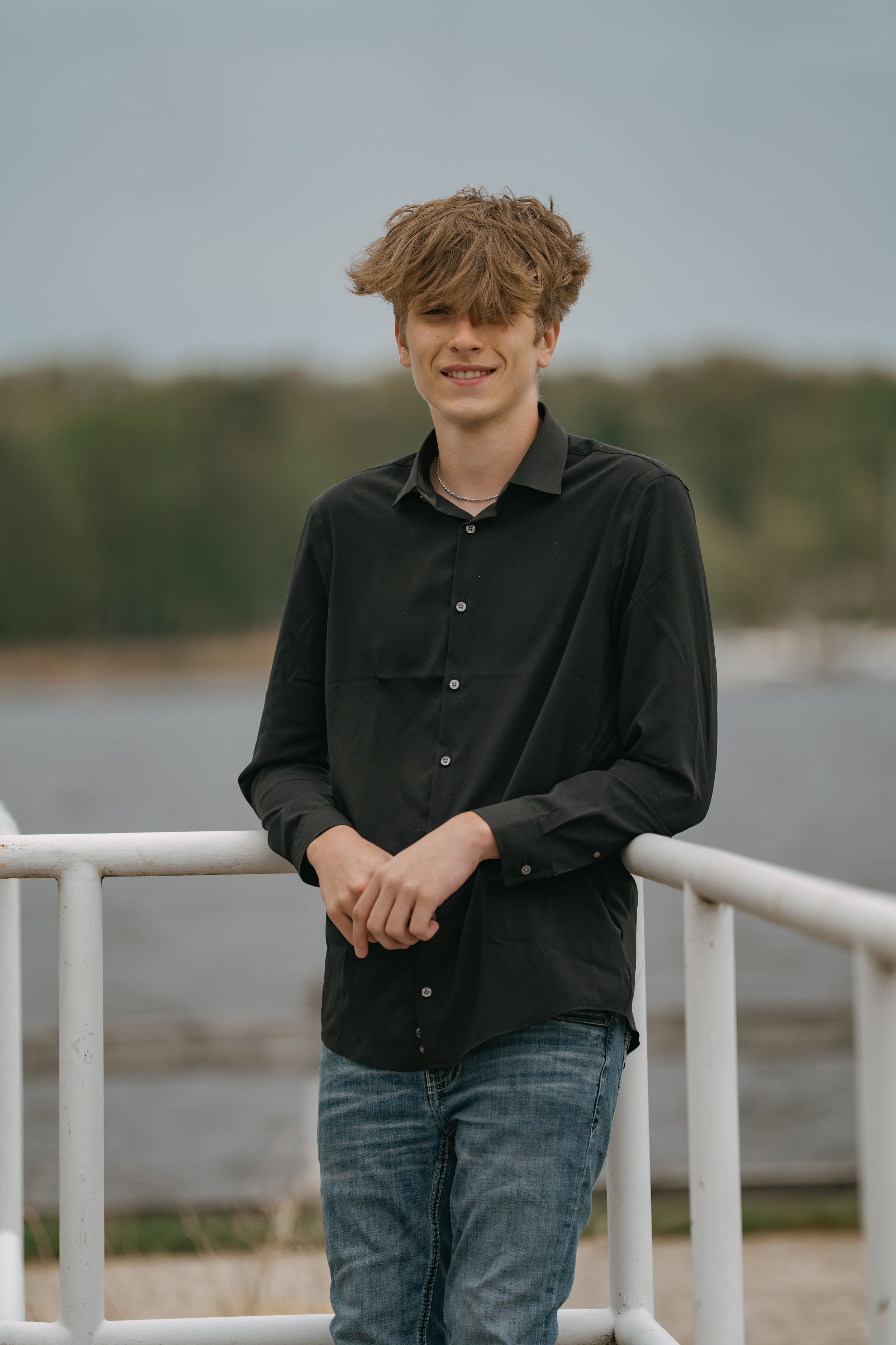Casual portrait of person in dark shirt standing on dock railing with water and trees in background.