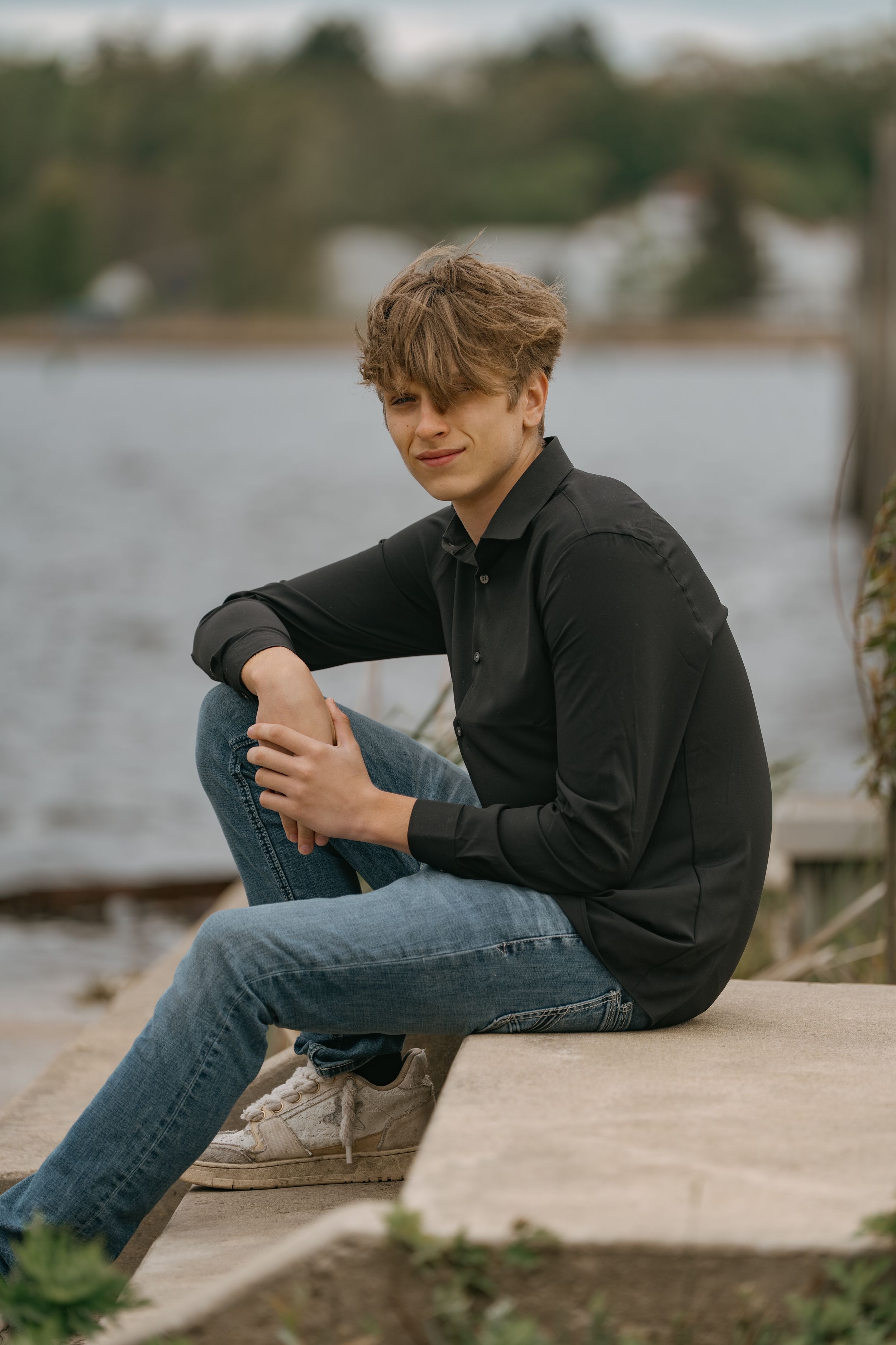 Someone in dark clothing sitting casually on a wooden dock by a lake.
