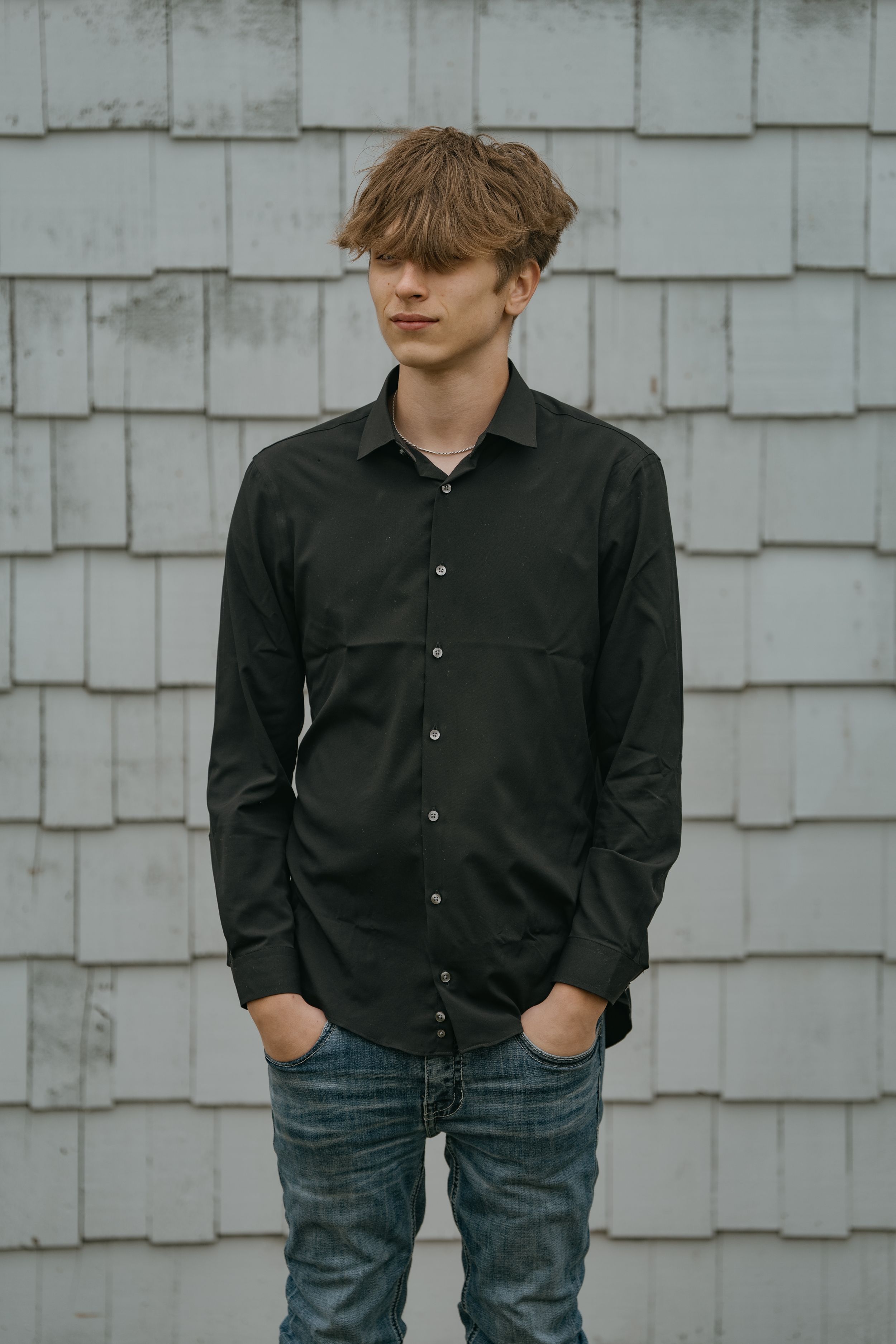 Portrait against white shingle wall showing someone in a dark button-down shirt and jeans.