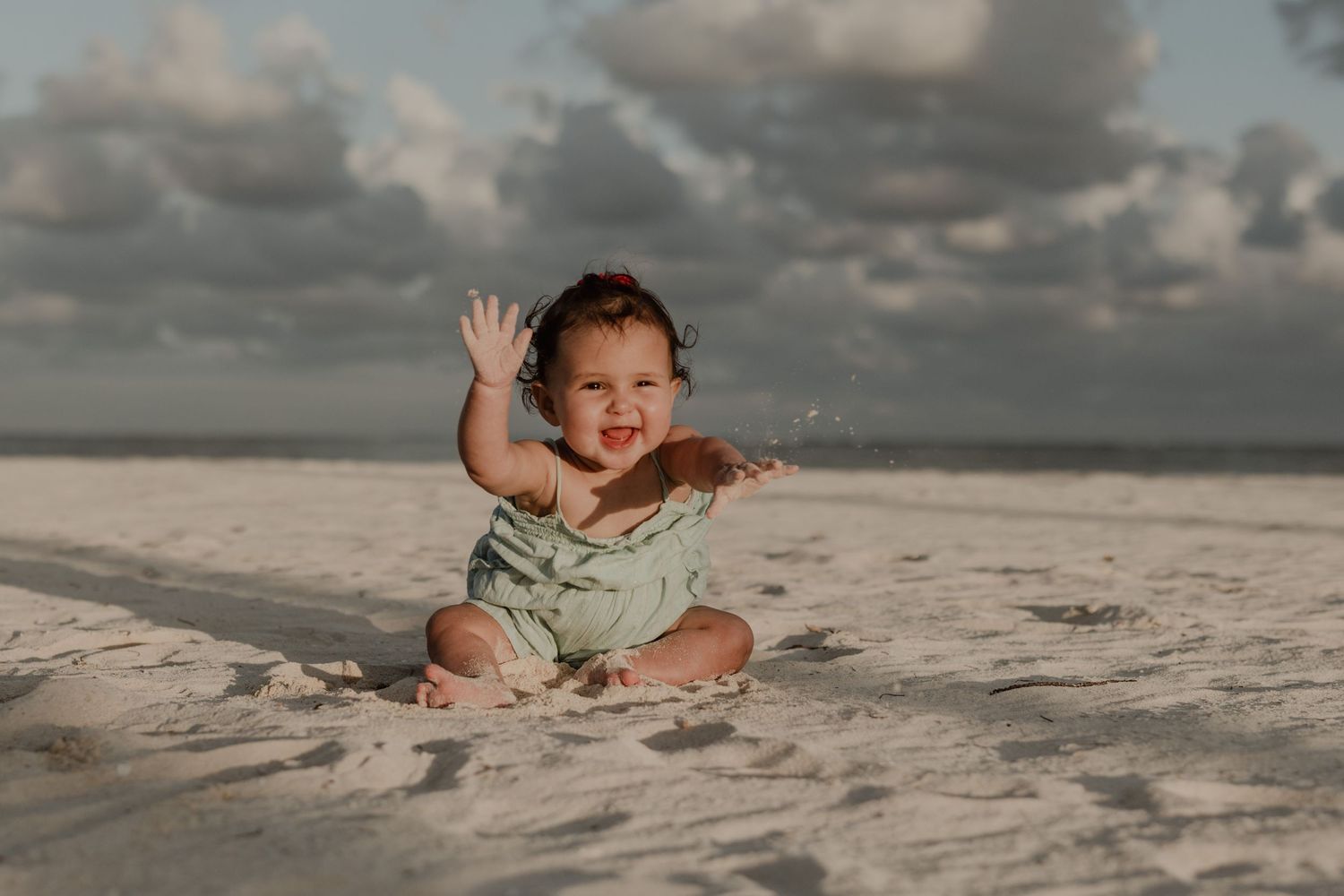Happy baby in green romper playing in white sand on a beach with cloudy sky backdrop.
