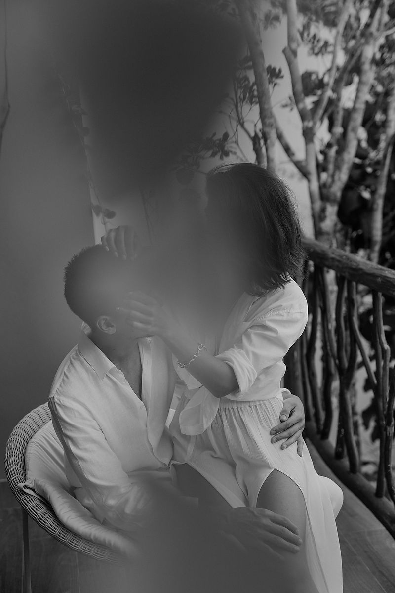 Black and white romantic photo of two people in white clothing embracing on a balcony with tree branches in background.