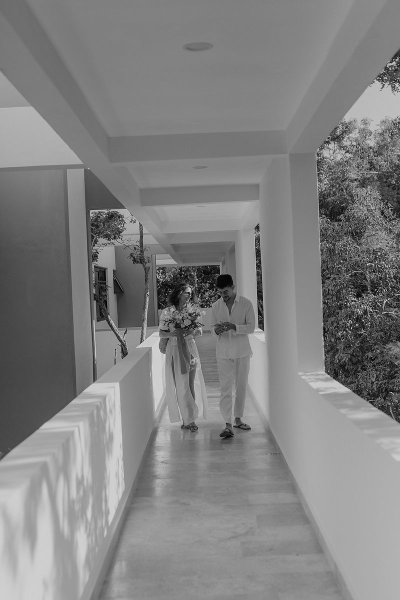 Two figures in light colored clothing stroll down a sunny outdoor corridor with white columns and walls.