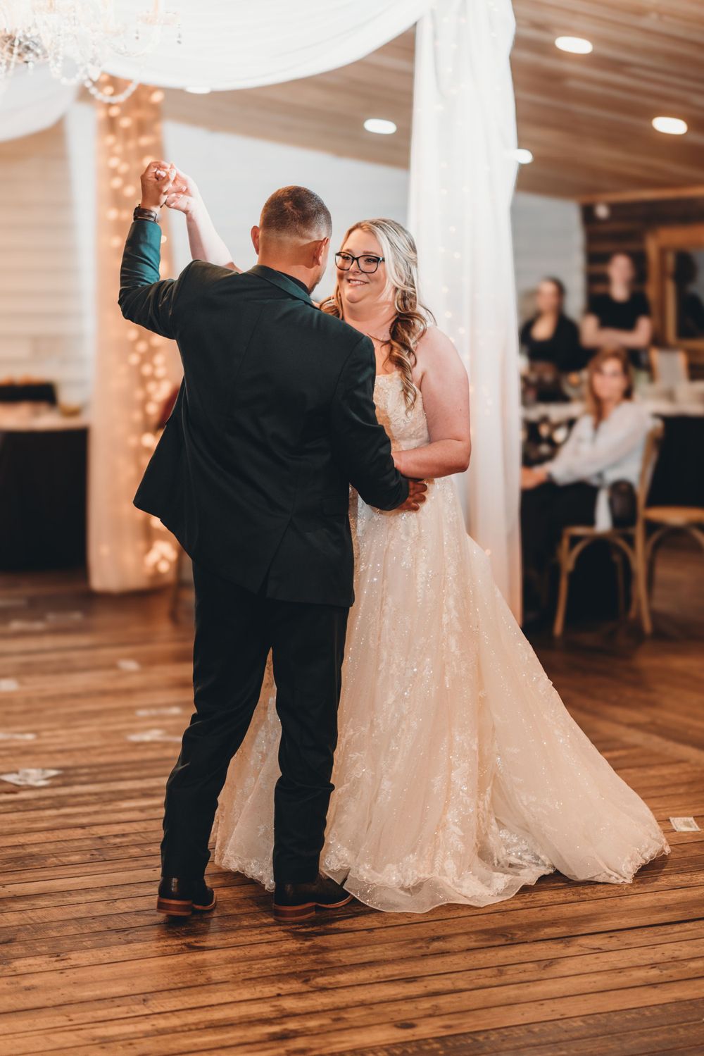 Couple shares intimate dance moment on wooden floor under draped fabric and chandelier.