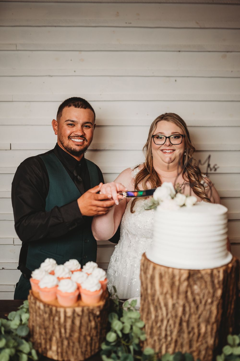 Wedding couple poses playfully at rustic cake display with cupcakes on wood slice stands.