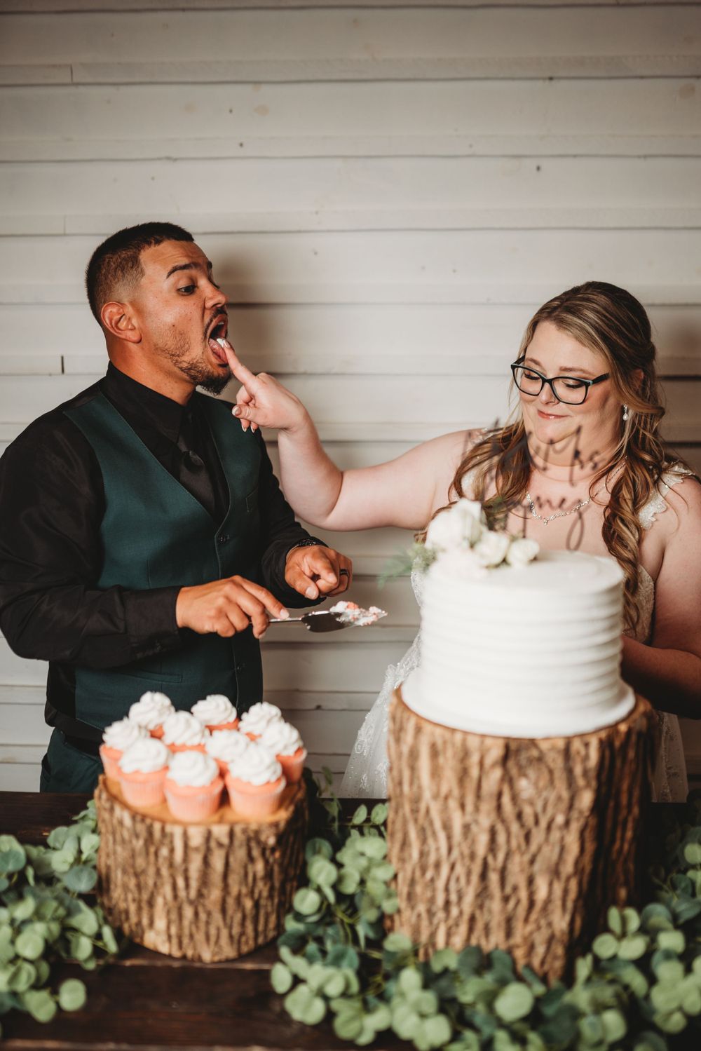 Newlyweds share playful cake-feeding moment at rustic-themed wedding dessert display.