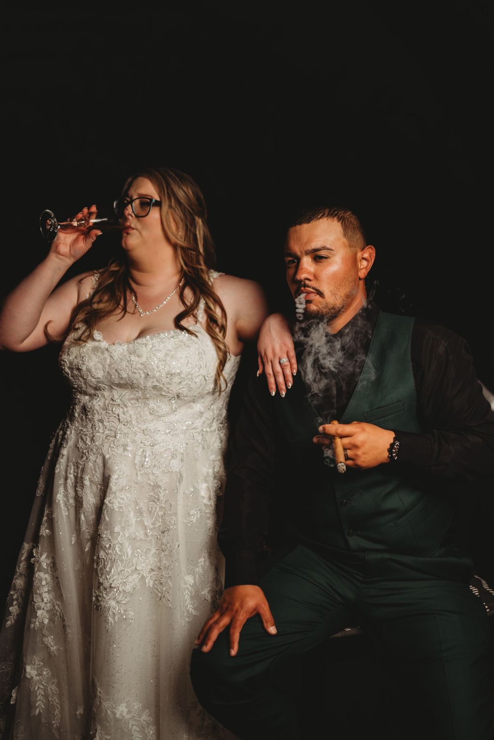 Newlyweds share a playful moment smoking cigars in formal wedding attire against a dark background.