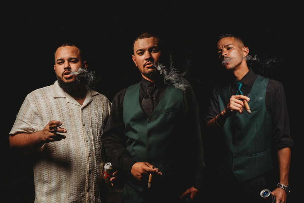 Three men in formal attire posing together while smoking cigars at an event.