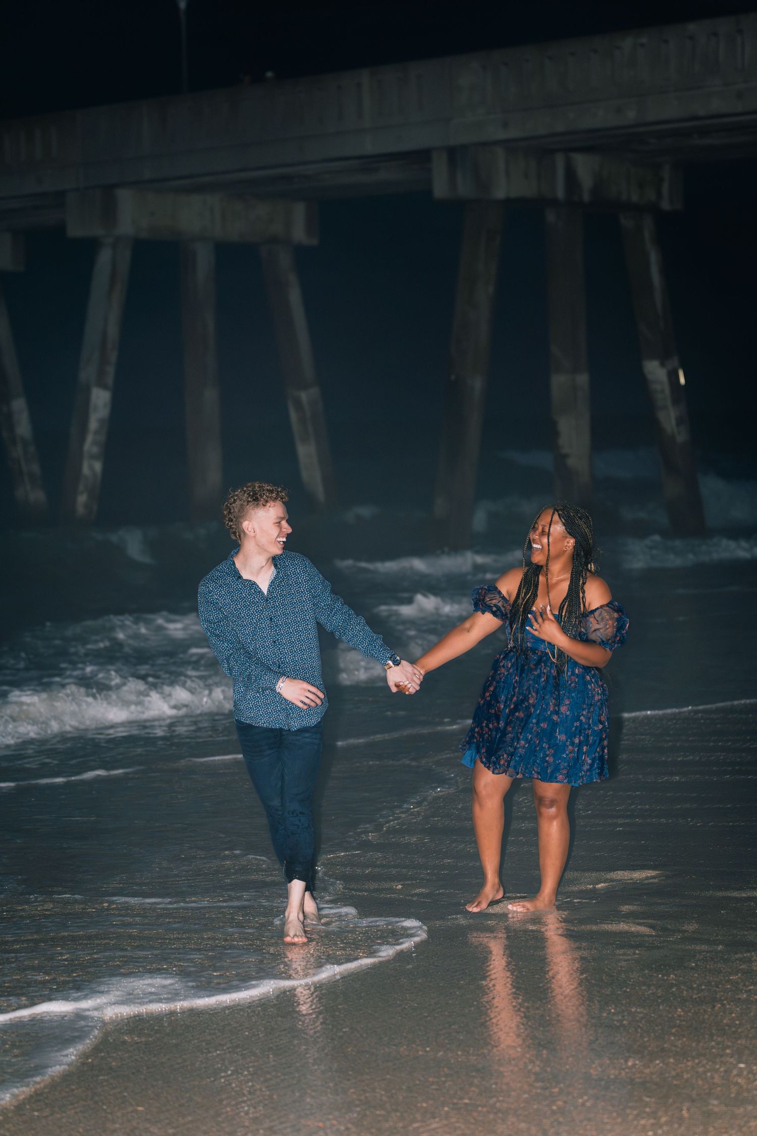 A couple holds hands while walking along the beach at night under a pier.