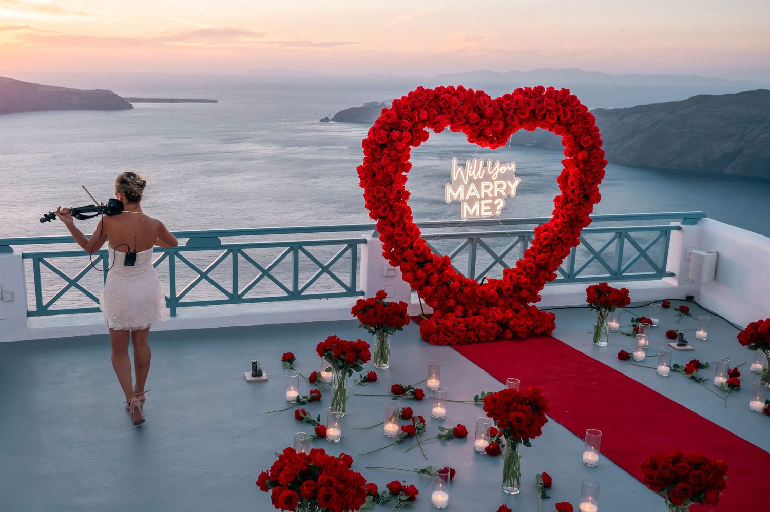 Romantic marriage proposal setup with red rose heart arch and 'Marry Me' sign overlooking Santorini sunset.