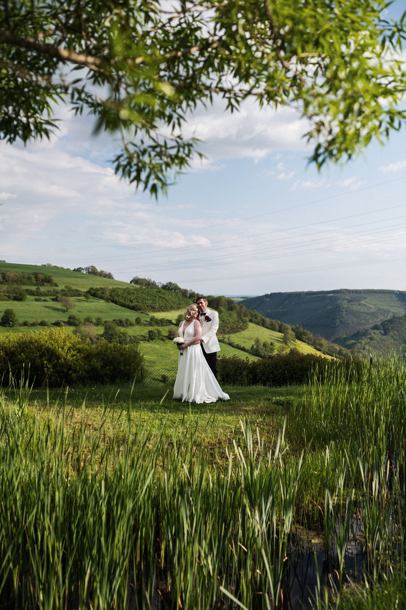 A couple embraces in a romantic pose on a hilltop meadow overlooking rolling green valleys on a sunny afternoon.