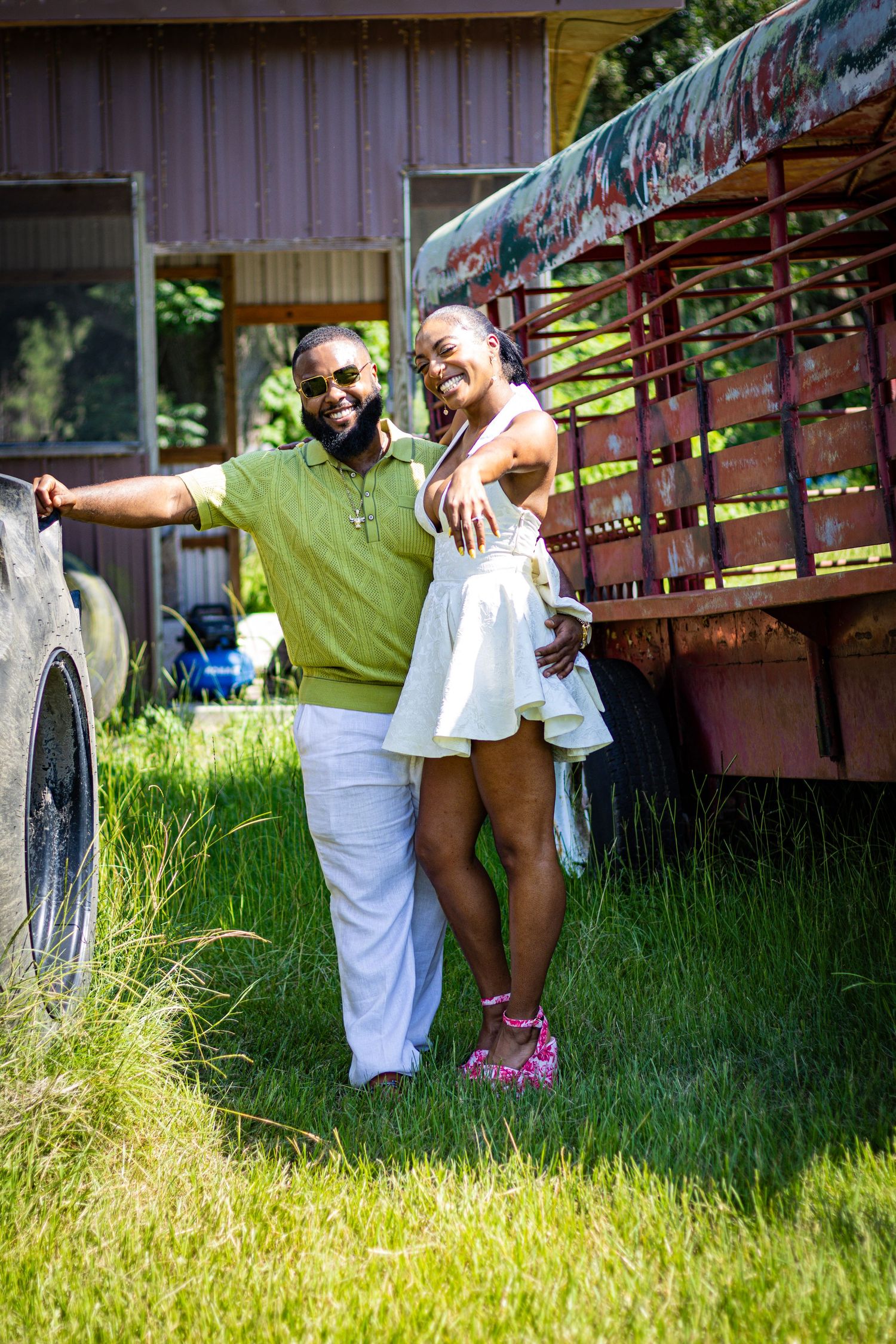 Couple posing playfully by rustic trailer in sunny outdoor setting.
