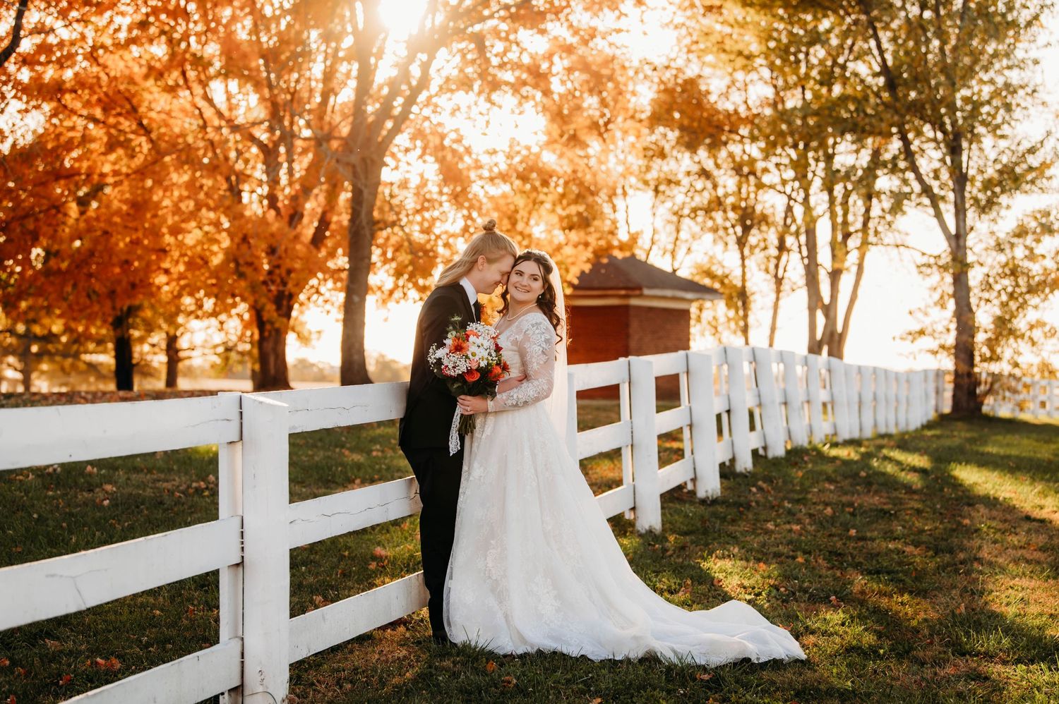 A sunset wedding portrait along a white fence with fall foliage in the background.