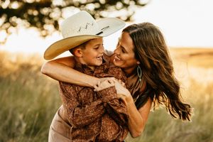 A couple in western attire embraces during a golden hour photoshoot in a grassy field.