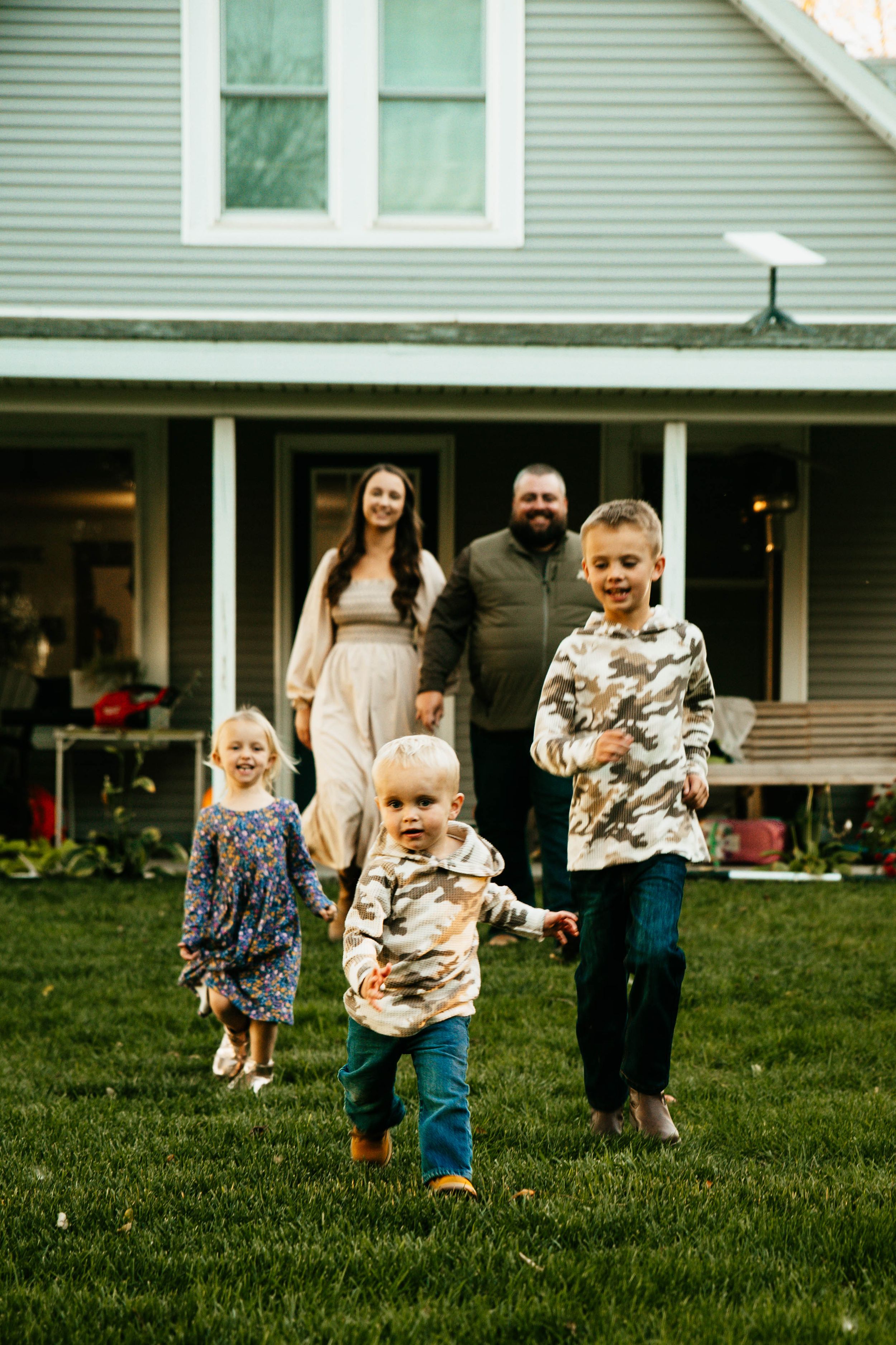 Children run and play together on a green lawn in front of a gray house on a summer day.