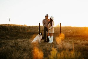 A couple embraces in a golden field during sunset while a photographer captures their romantic moment.