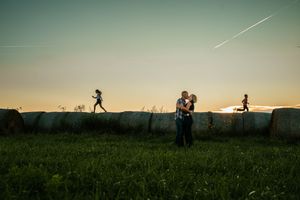 Silhouettes of people in a grassy field at sunset with hay bales and a contrail in the sky.