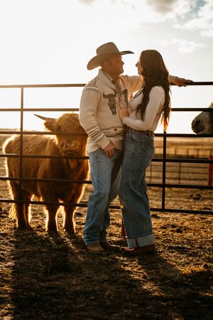 A couple in western attire shares a romantic moment against a rustic ranch fence at golden hour.