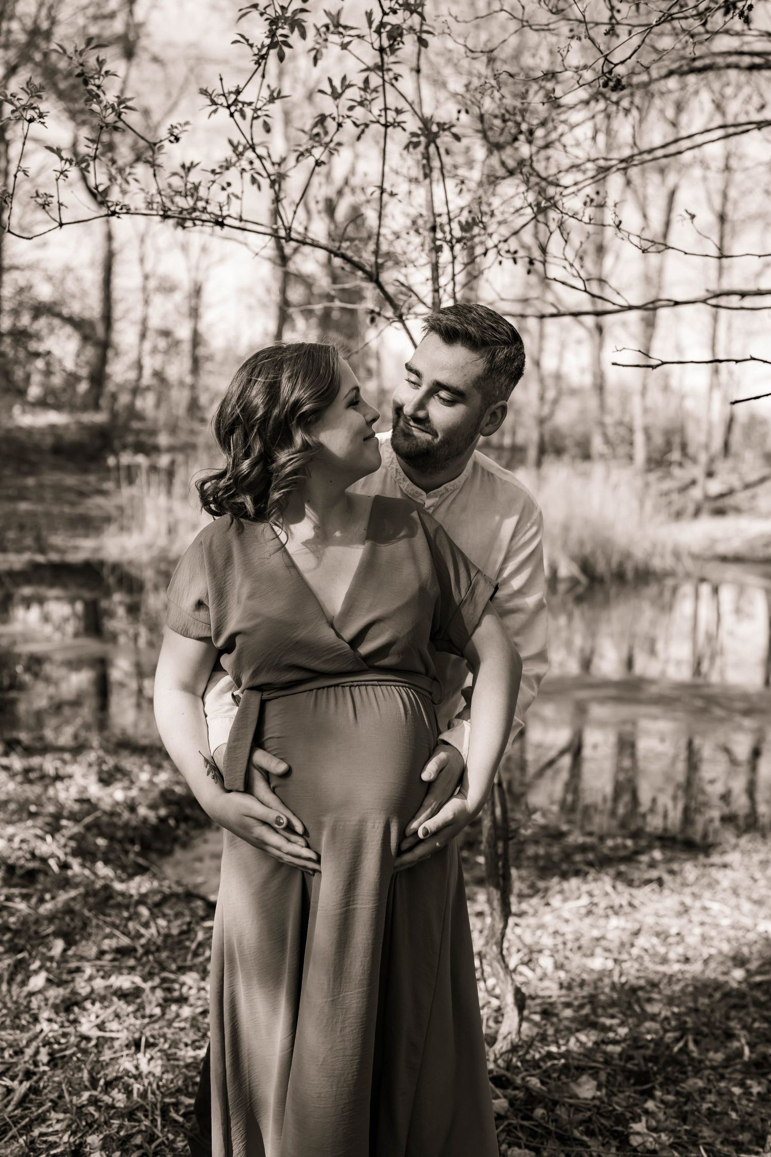 Romantic black and white maternity photo of couple embracing by pond in wooded setting.