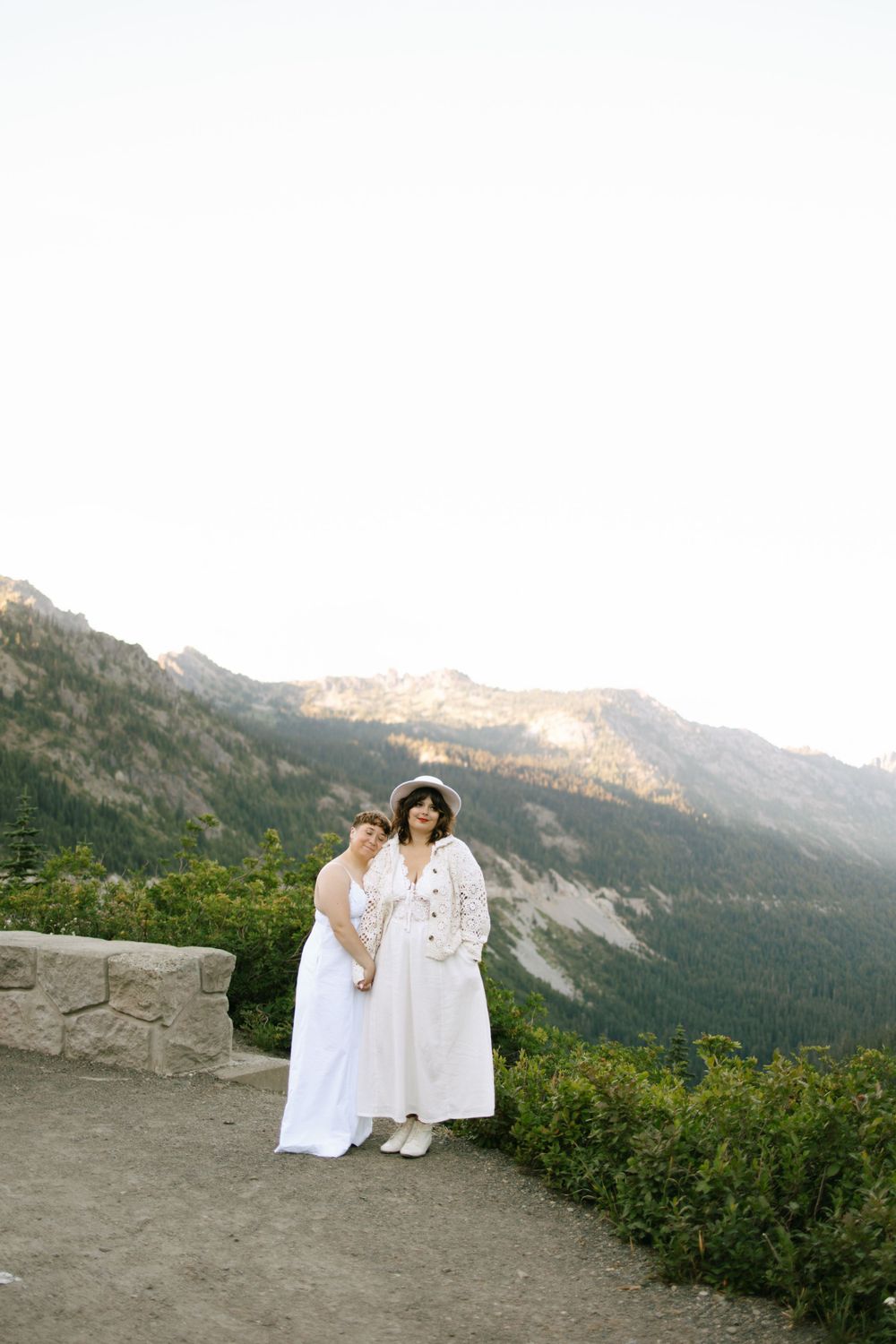 Two brides in white dresses posing together at mountain overlook at sunset.
